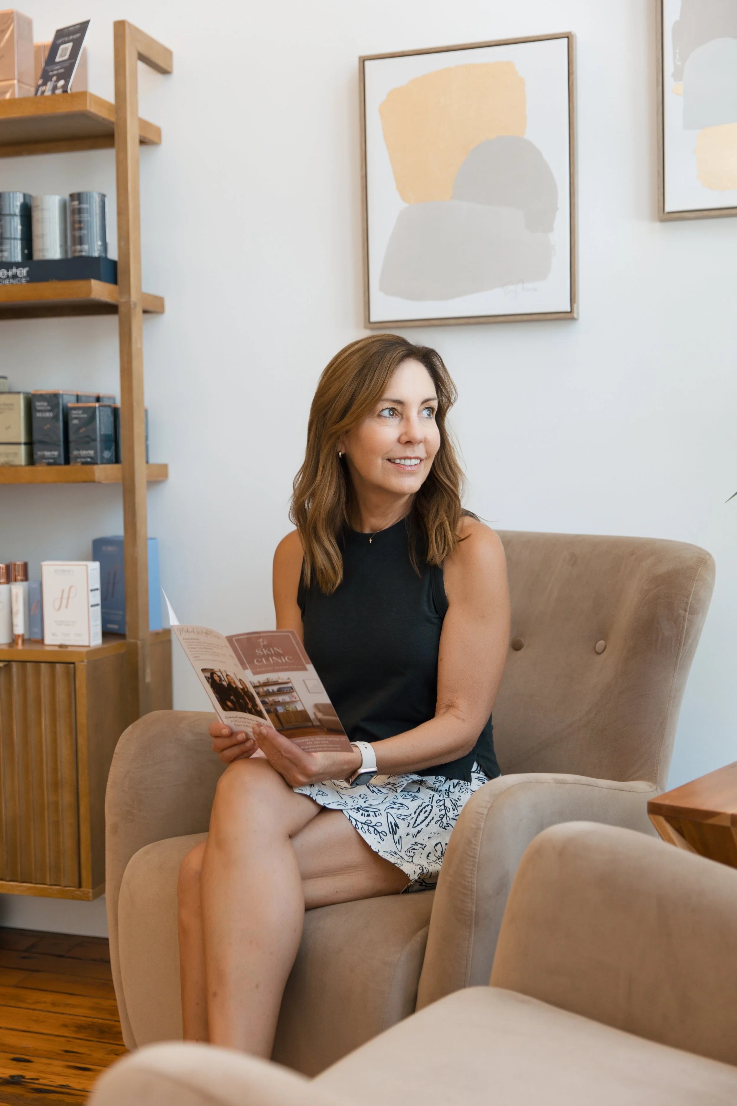 A woman sitting in a beige armchair holding a brochure, in a room with abstract artwork and a wooden shelf with products.