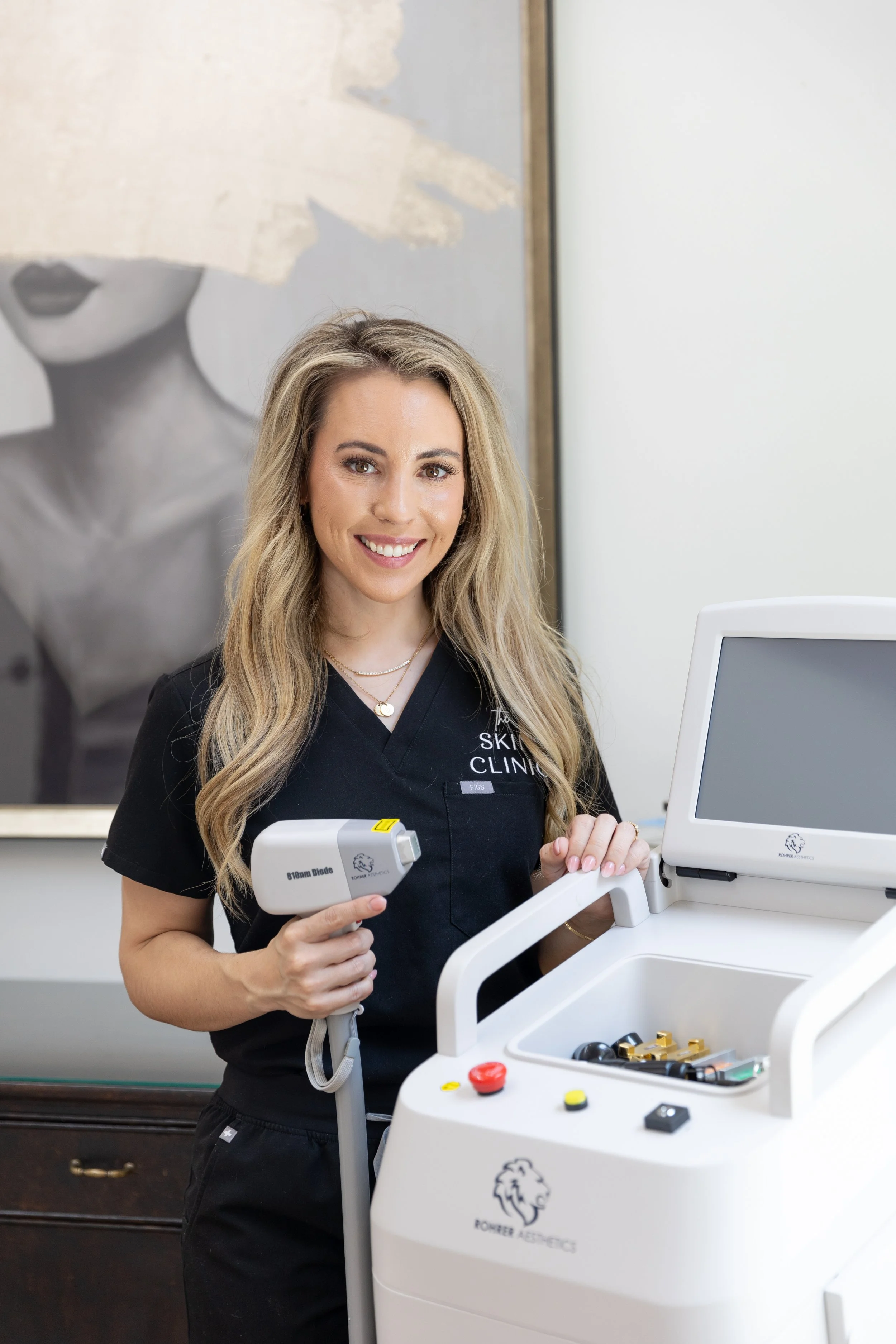A woman with long blonde hair smiling in a medical clinic with laser equipment, wearing black scrubs with the words "The Skin Clinic" on the chest, standing beside a white laser device.