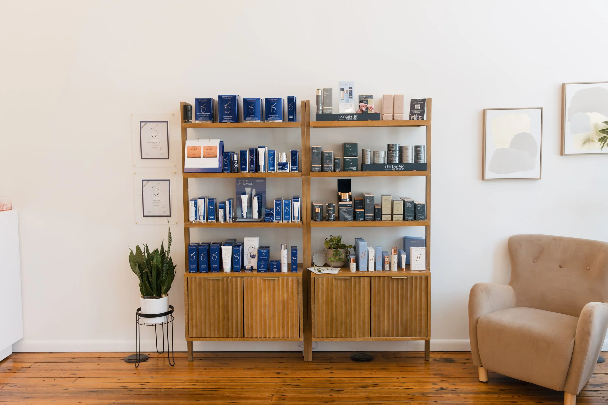 A wooden display shelf filled with skincare products, handbags, and skincare tools, a potted plant on a stand, and a beige armchair in a well-lit room with wooden flooring.