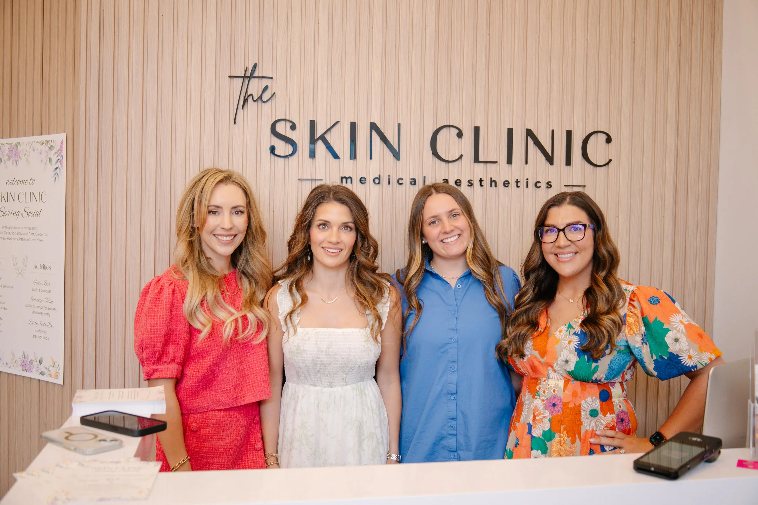 Four women standing behind a reception desk at a skin care clinic, smiling for the photo.