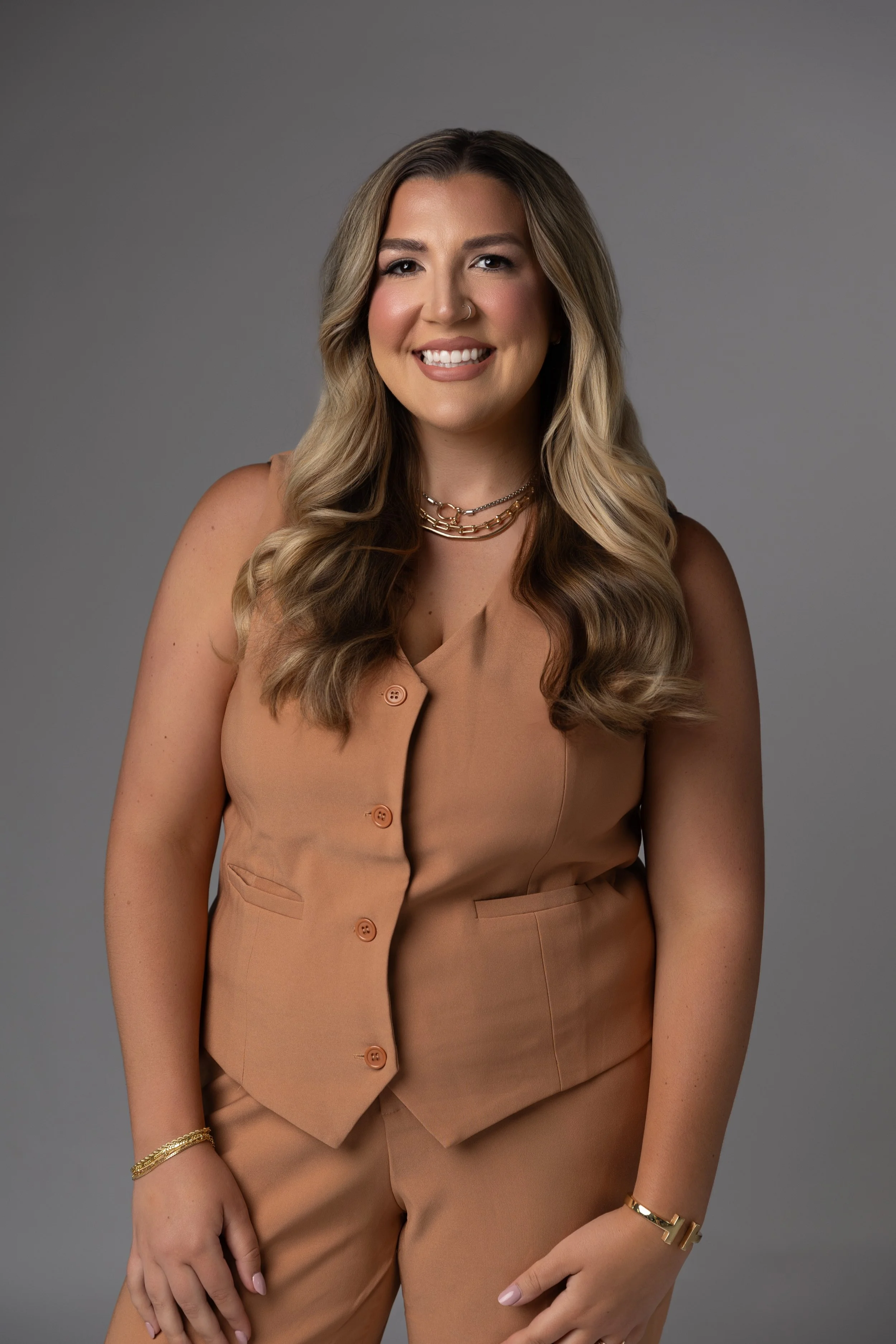 A smiling woman with wavy blonde hair, wearing a tan sleeveless vest and matching pants, accessorized with layered necklaces and bracelets, posing against a plain gray background.