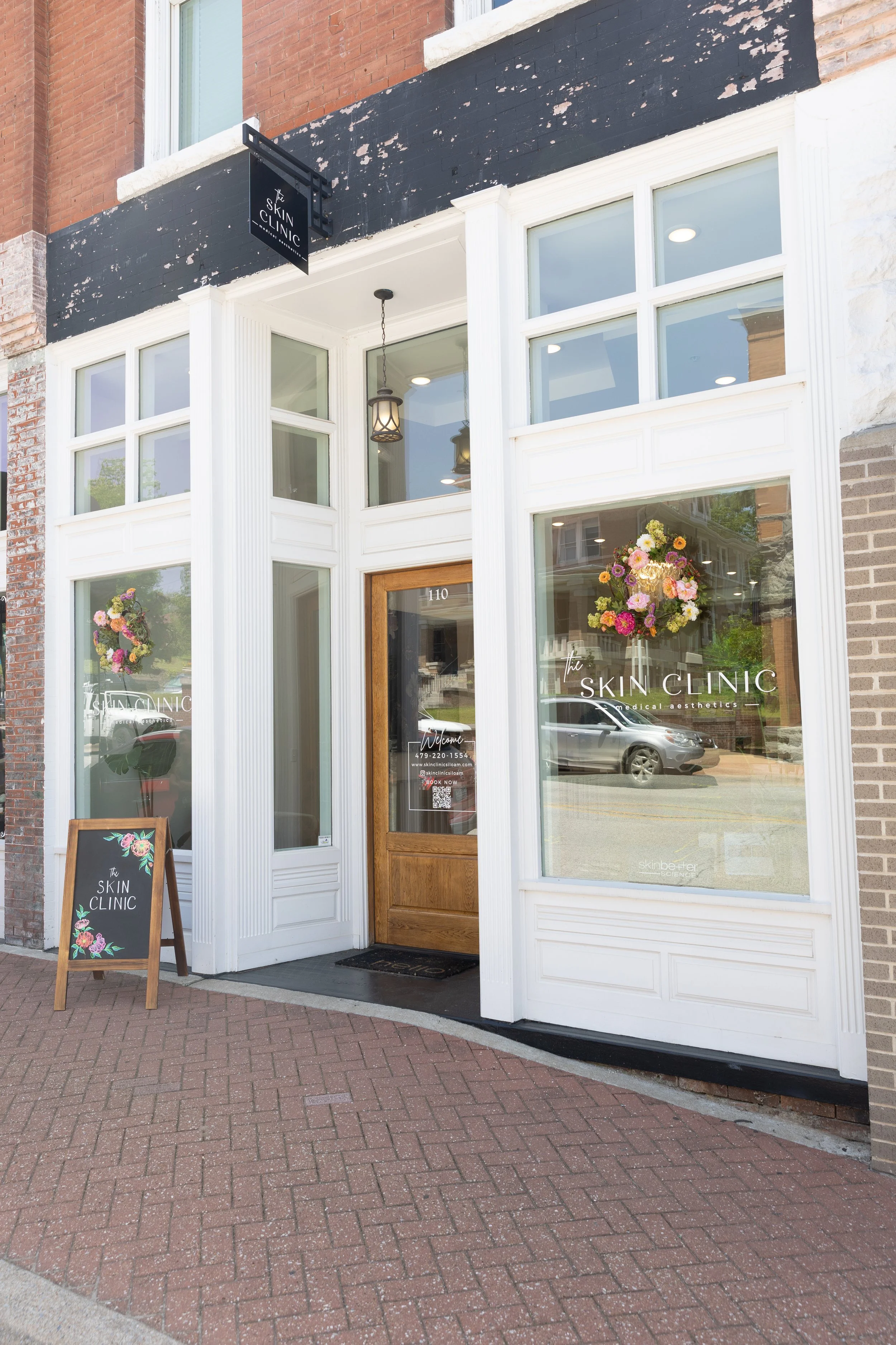 Exterior view of a storefront for The Skin Clinic, with large glass windows, a wooden door, flower arrangements, and a sidewalk with brick paving.