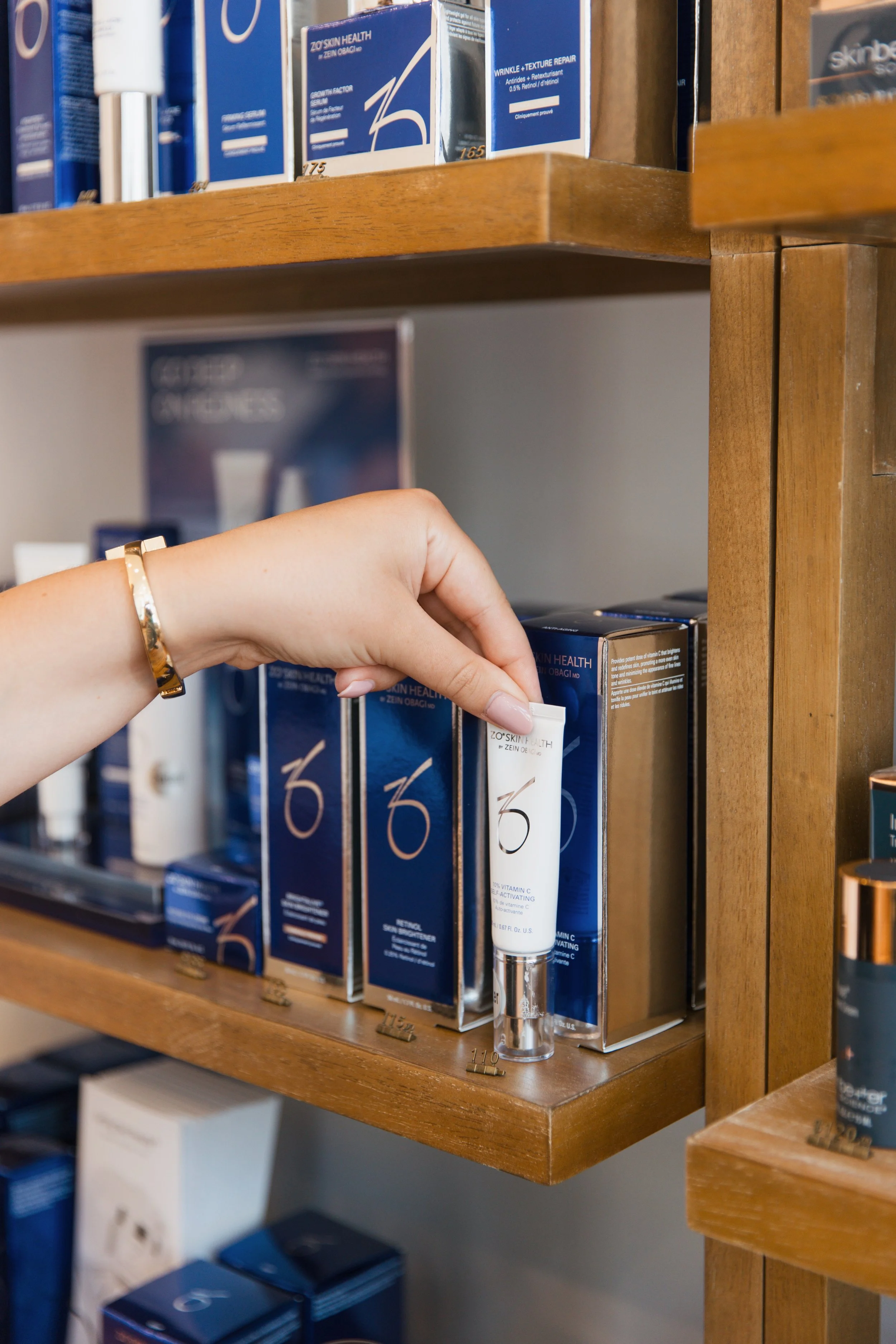 A person with a gold bracelet picking up a skincare product from a wooden shelf, shelves stocked with various skincare products and boxes.