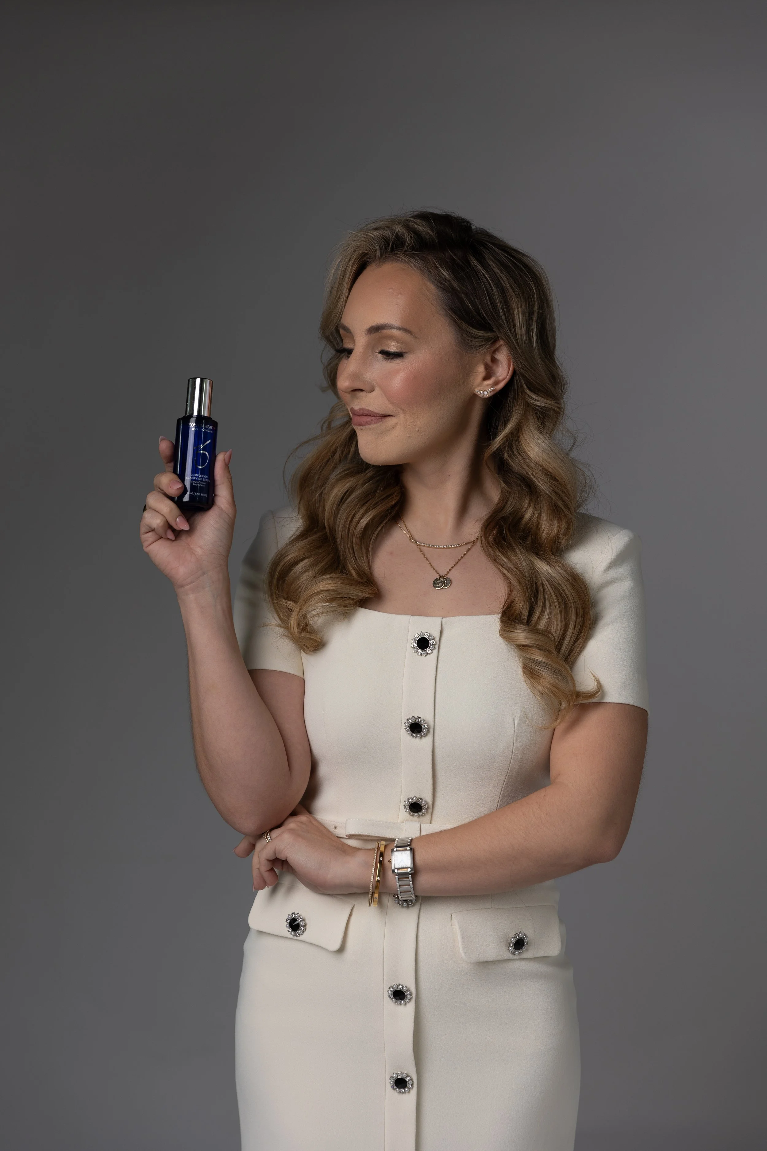 Woman with wavy hair dressed in a cream-colored dress holding a small dark blue bottle, looking at it with a slight smile.