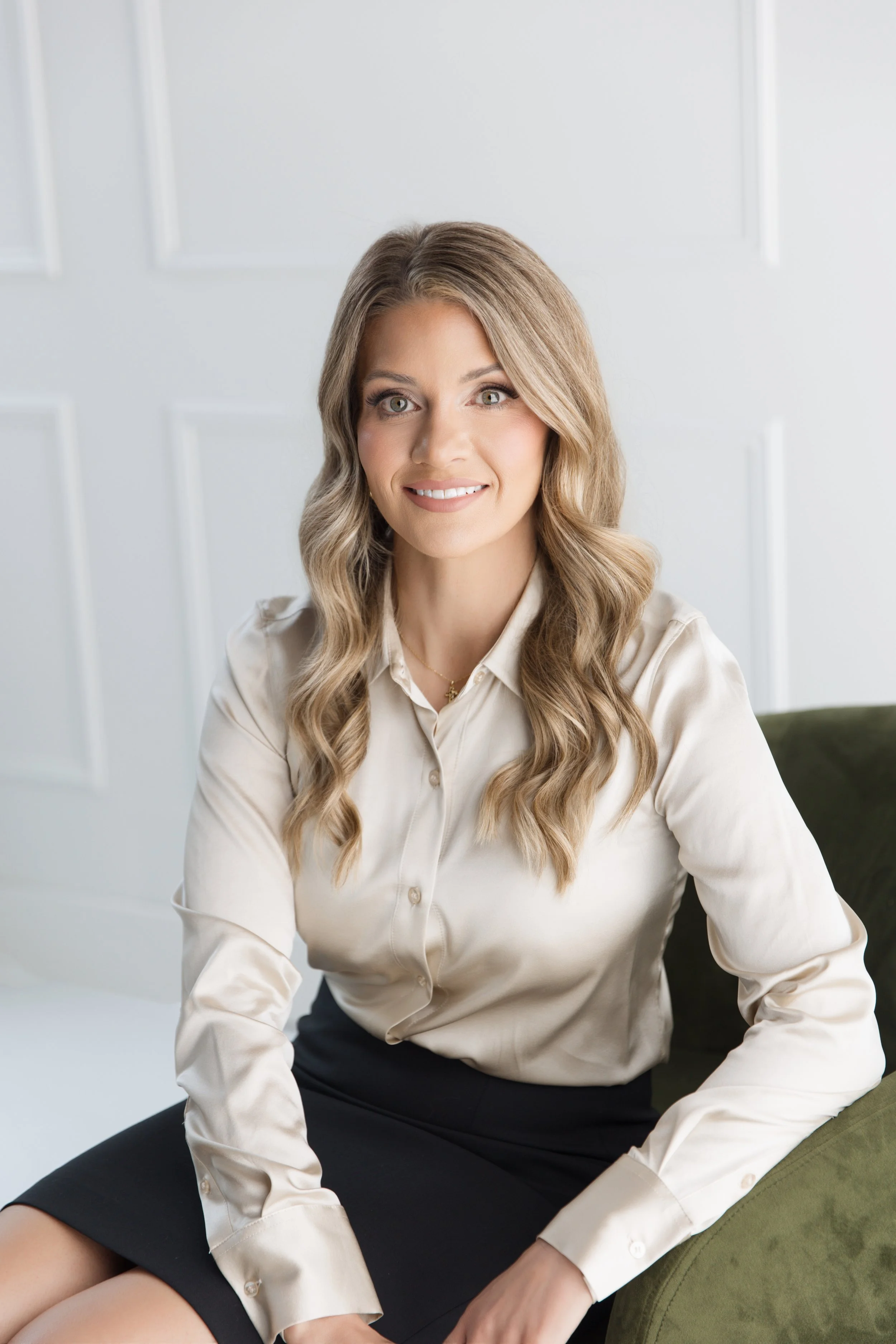 A woman with long wavy blonde hair, wearing a cream-colored blouse and a black skirt, sitting on a green armchair in front of a white wall with decorative molding, smiling at the camera.