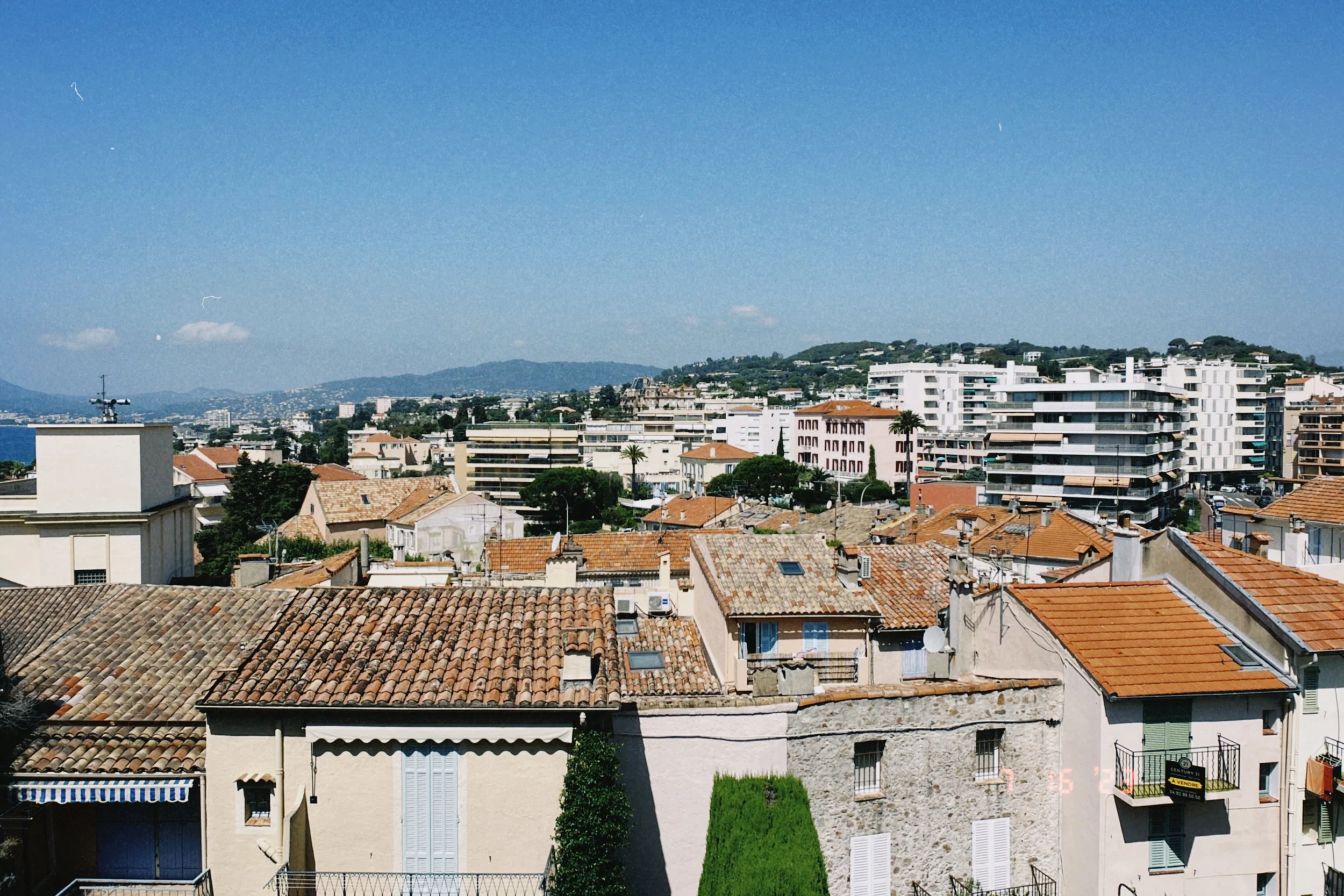Cityscape with a mix of old and modern buildings, terracotta rooftops, green trees, hills in the background, and a clear blue sky.