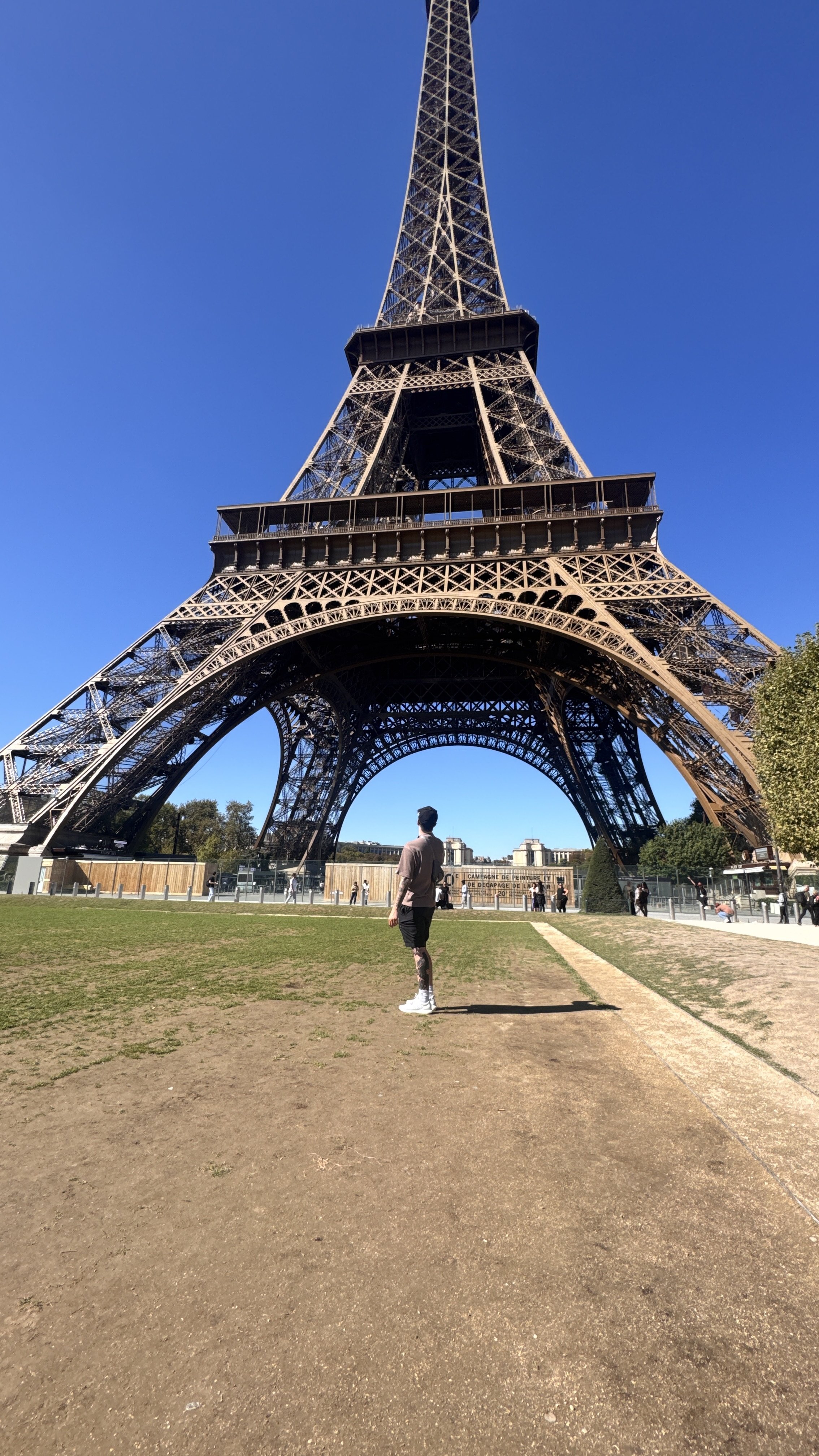 A person standing on a dirt path in front of the Eiffel Tower in Paris, France, under a clear blue sky.