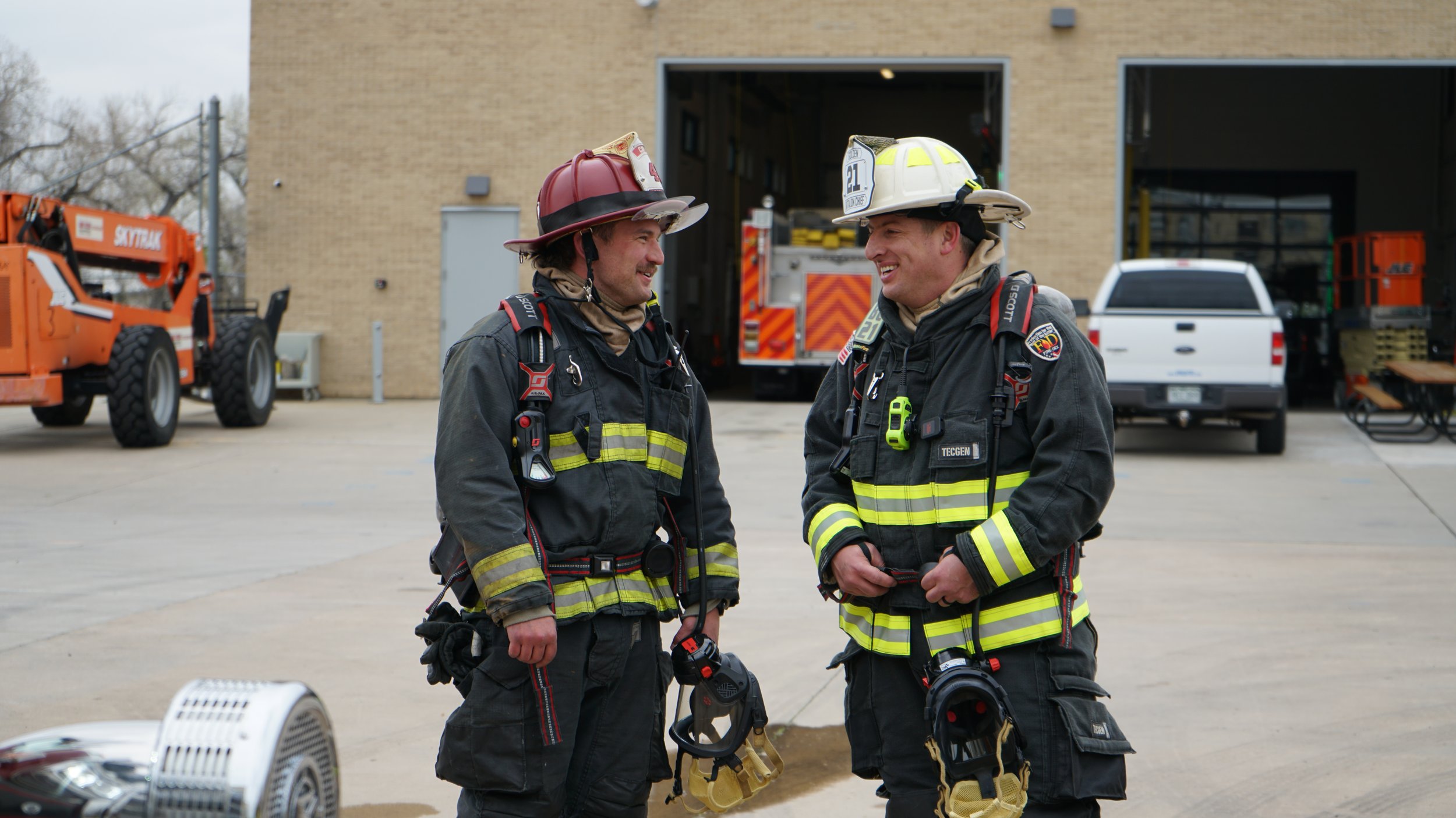 Two male firefighters in black turnout gear and helmets standing outside in front of fire trucks, smiling and talking