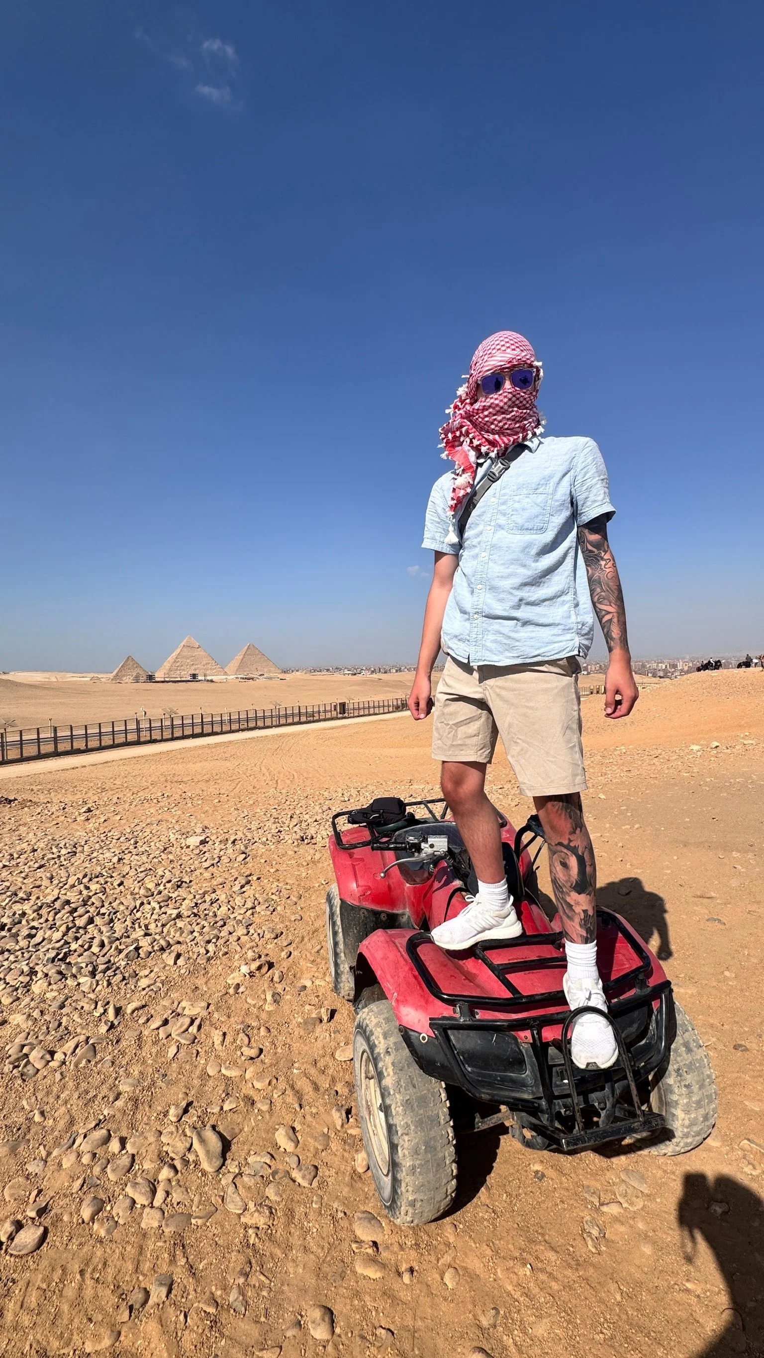 Person standing on a red ATV in the desert near the Pyramids of Giza under a clear blue sky, wearing sunglasses, a scarf over their face, and casual summer clothes.
