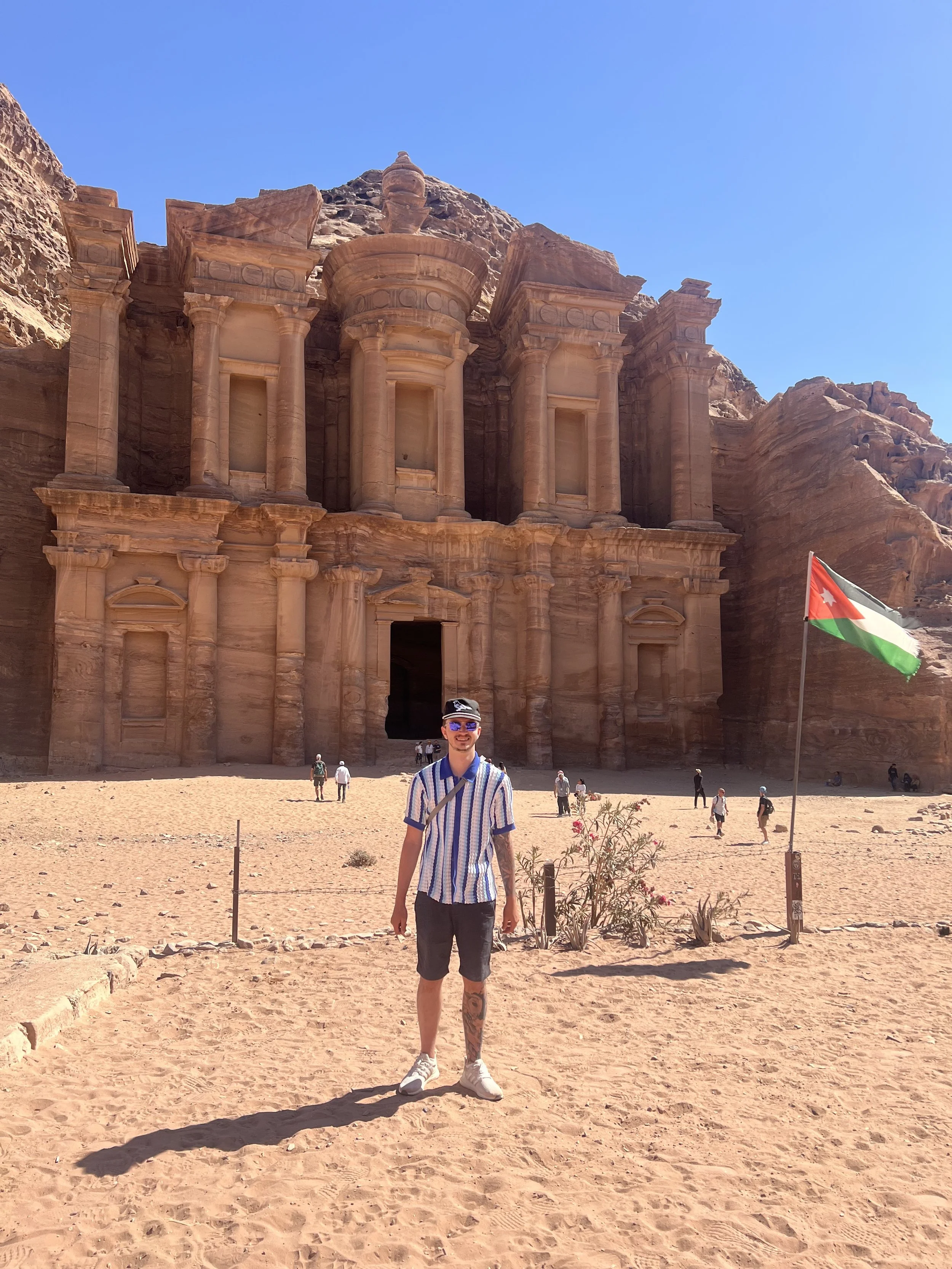 A man in a striped shirt and shorts standing in front of the ancient rock-cut city of Petra in Jordan, with large stone facades and cliffs behind him, alongside a Jordanian flag.