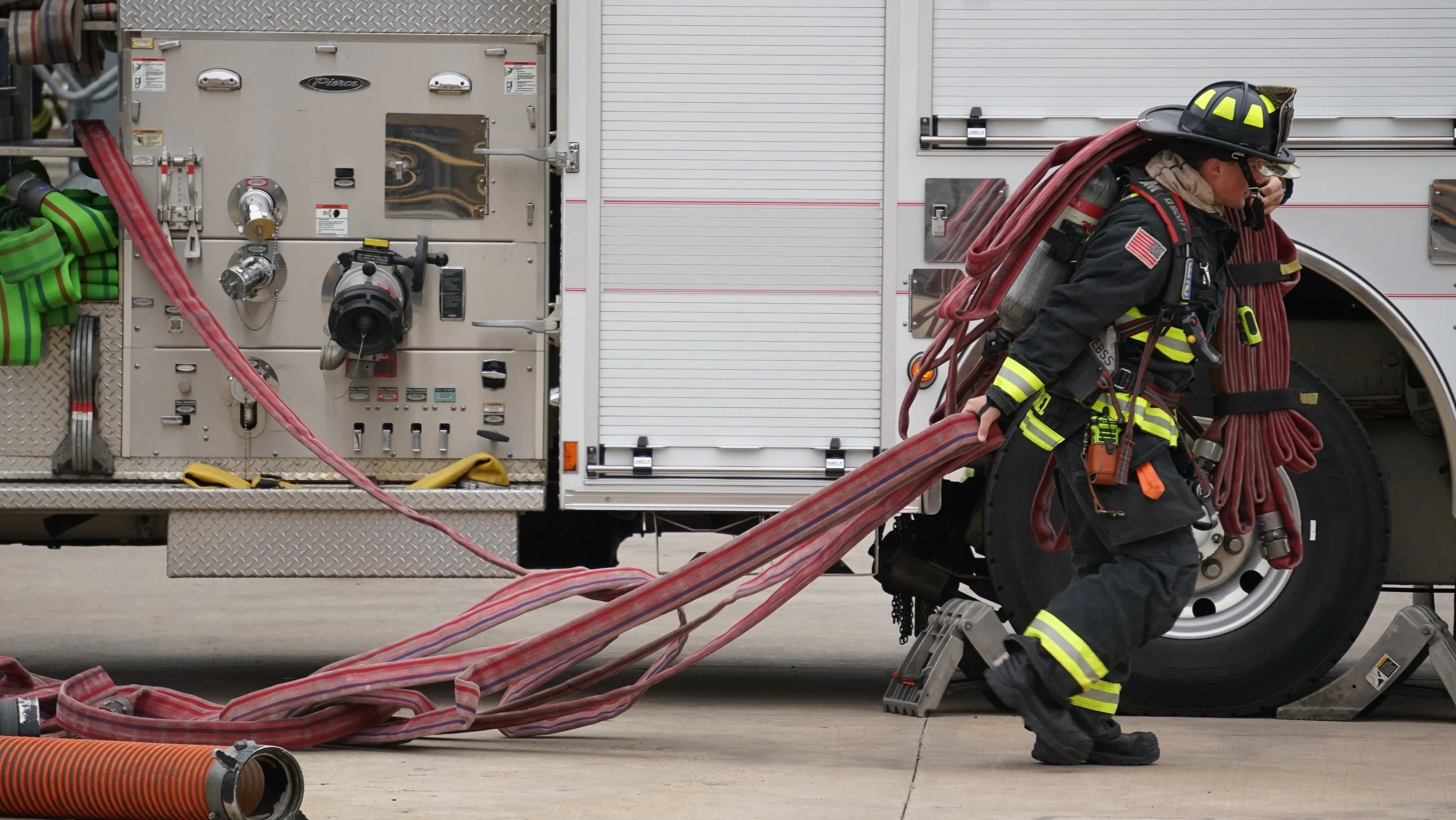A firefighter in black gear with yellow reflective stripes, wearing a helmet, is carrying fire hoses and equipment while walking next to a fire truck.