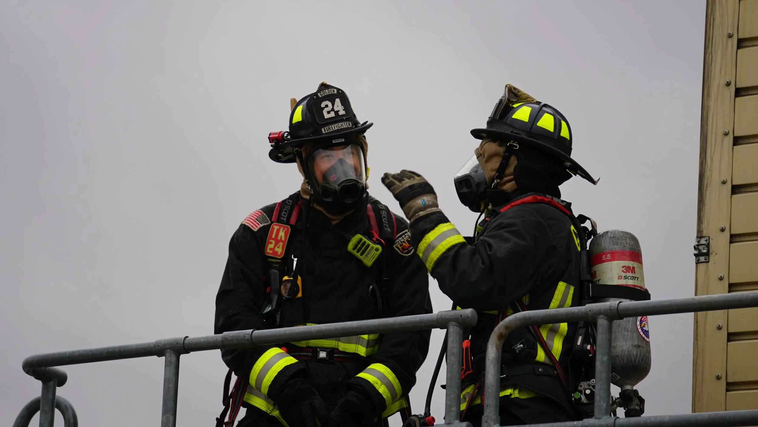 Two firefighters wearing full gear and helmets on a fire truck, engaged in conversation against a gray sky.