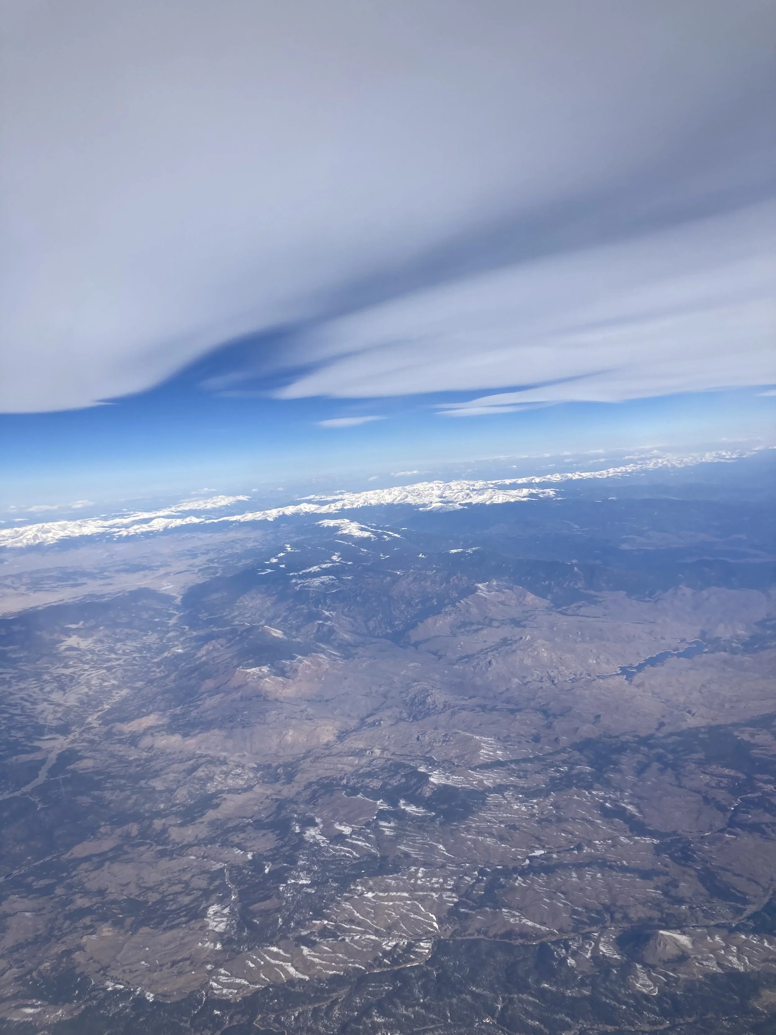 Aerial view of mountainous terrain with snow-capped peaks under a sky with wispy clouds.