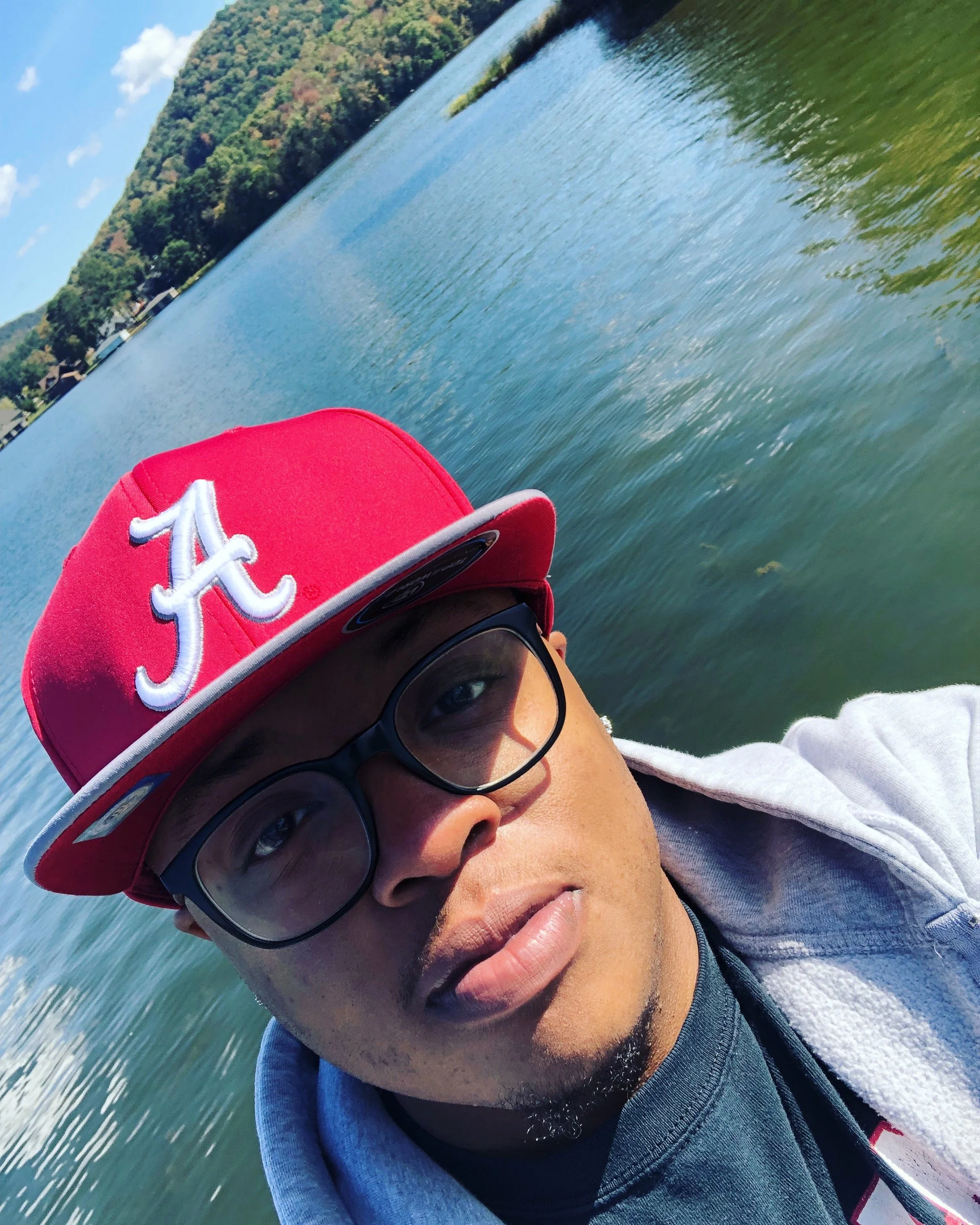 Close-up selfie of a young man wearing a red baseball cap with an 'A' logo, black glasses, and a gray hoodie, near a body of water with trees and a hill in the background.