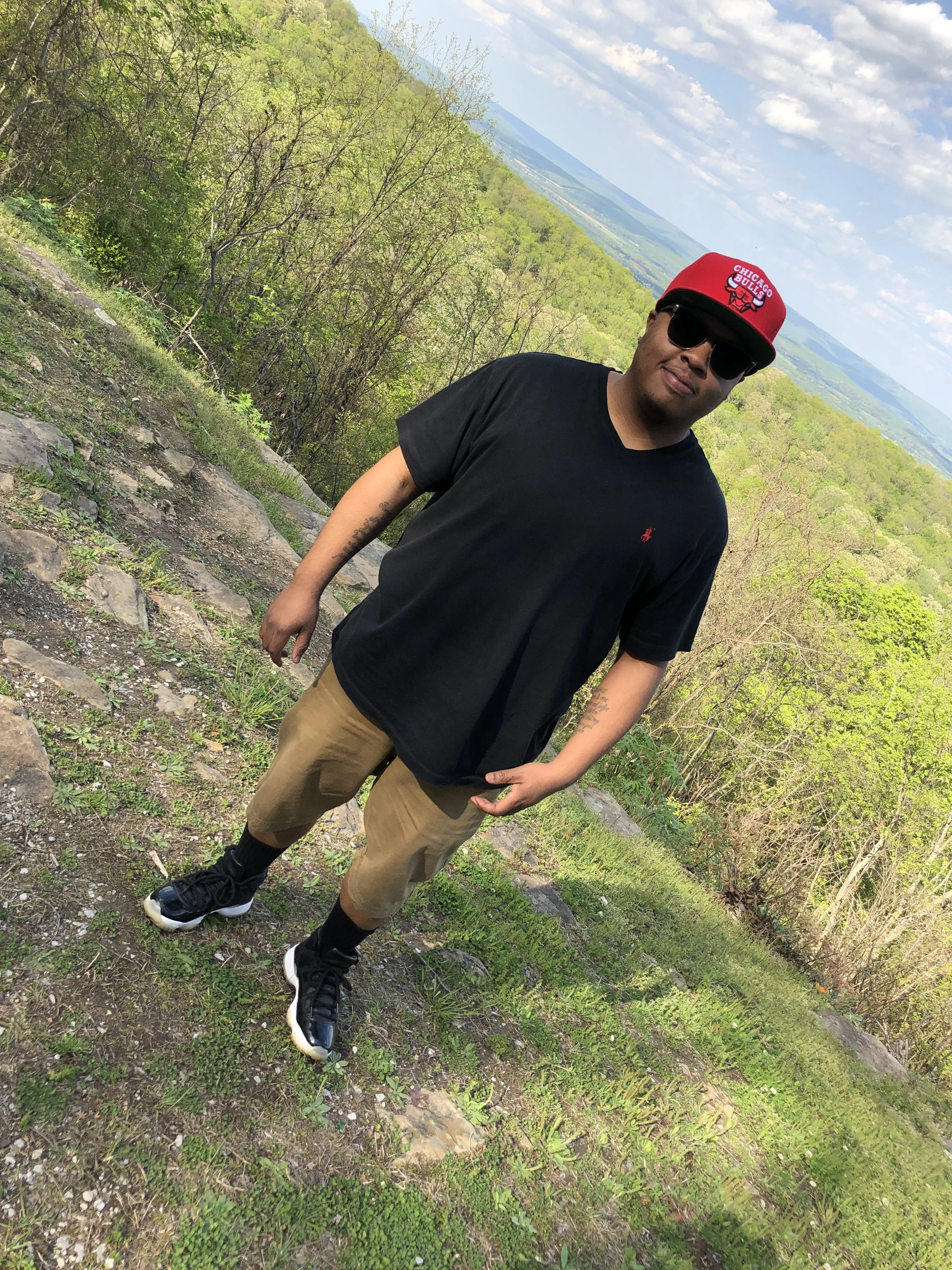A man standing outdoors on a hill with a view of lush green trees, mountains, and a partly cloudy sky in the background. He is wearing a red Chicago Bulls cap, black T-shirt, khaki shorts, black socks, and sneakers, and is wearing sunglasses.