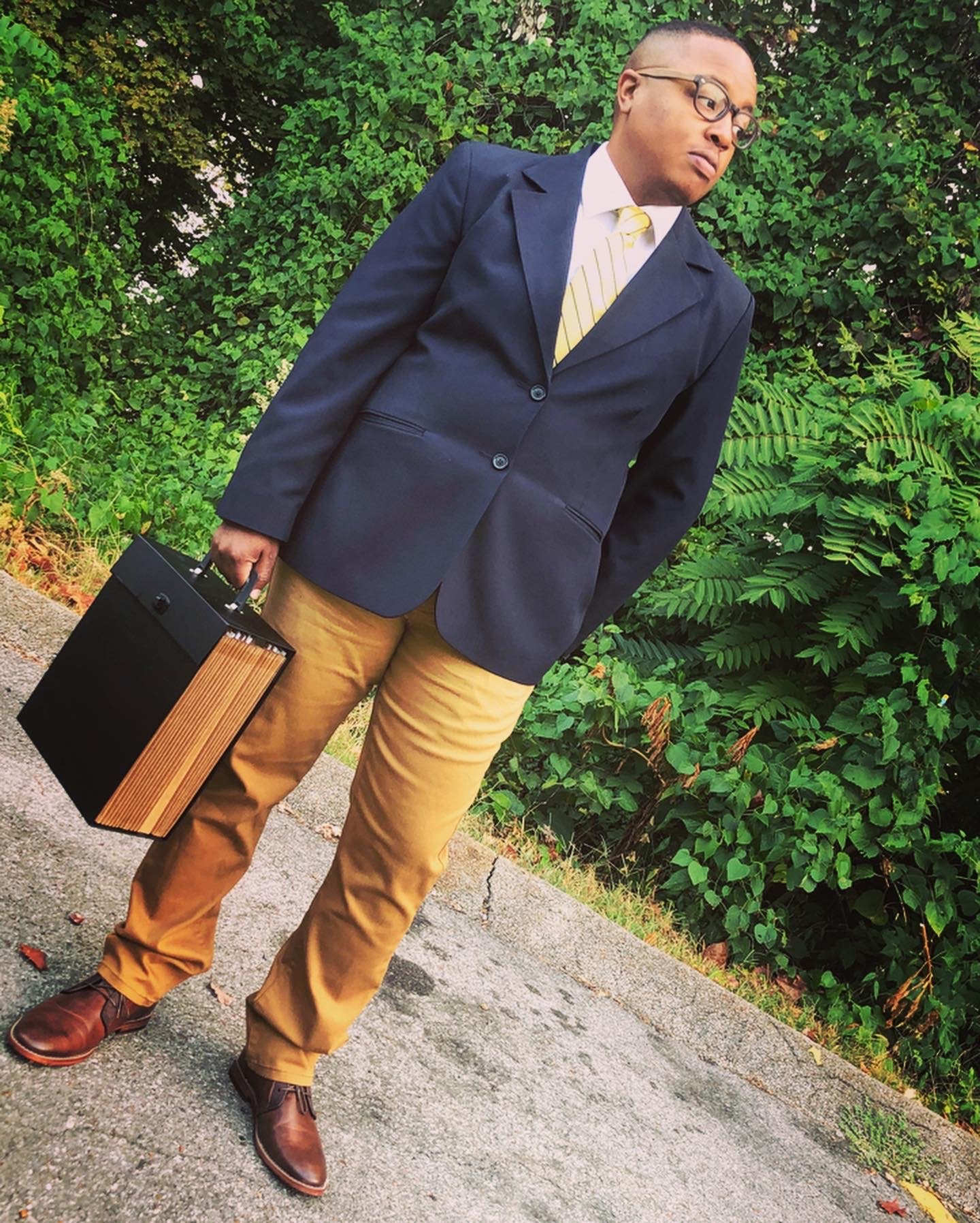 A man wearing glasses, a navy blazer, light yellow pants, and brown shoes, holding a black and wood-colored briefcase, standing outdoors against a green leafy background.