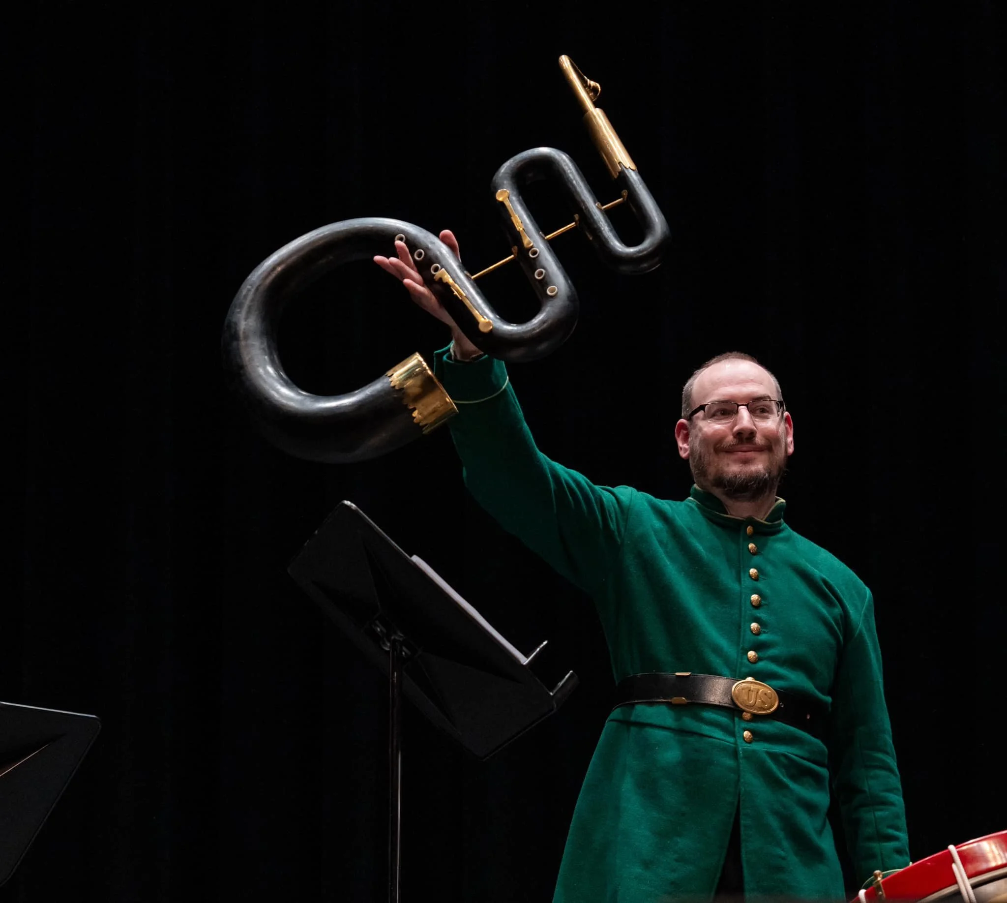 A man dressed in a green coat and a belt, holding a large, stylized black and gold instrument resembling a key with a musical note shape, against a dark background.