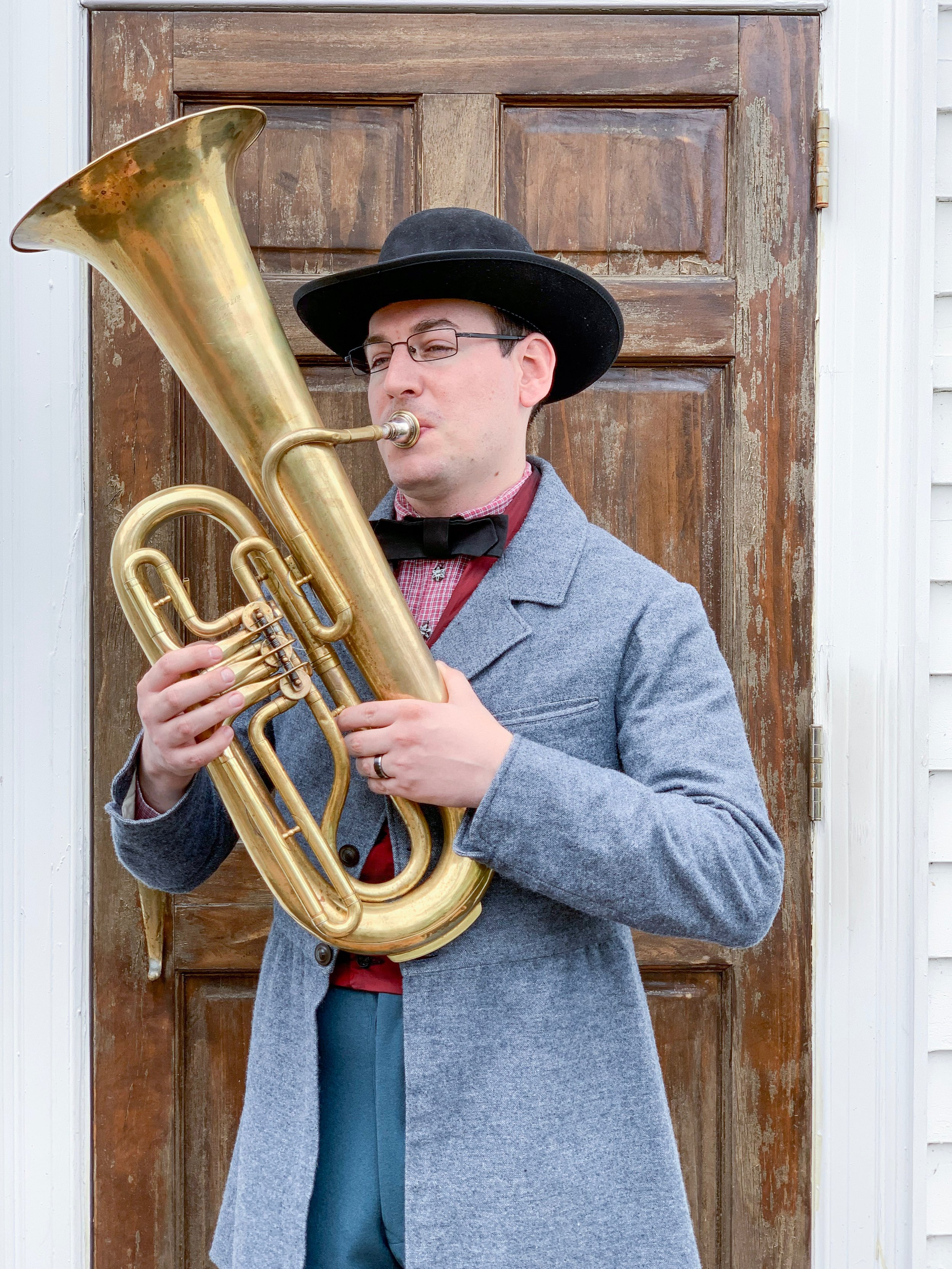 A man wearing glasses, a black hat, a bow tie, gray coat, and red shirt playing a brass tuba outdoors in front of a wooden door.