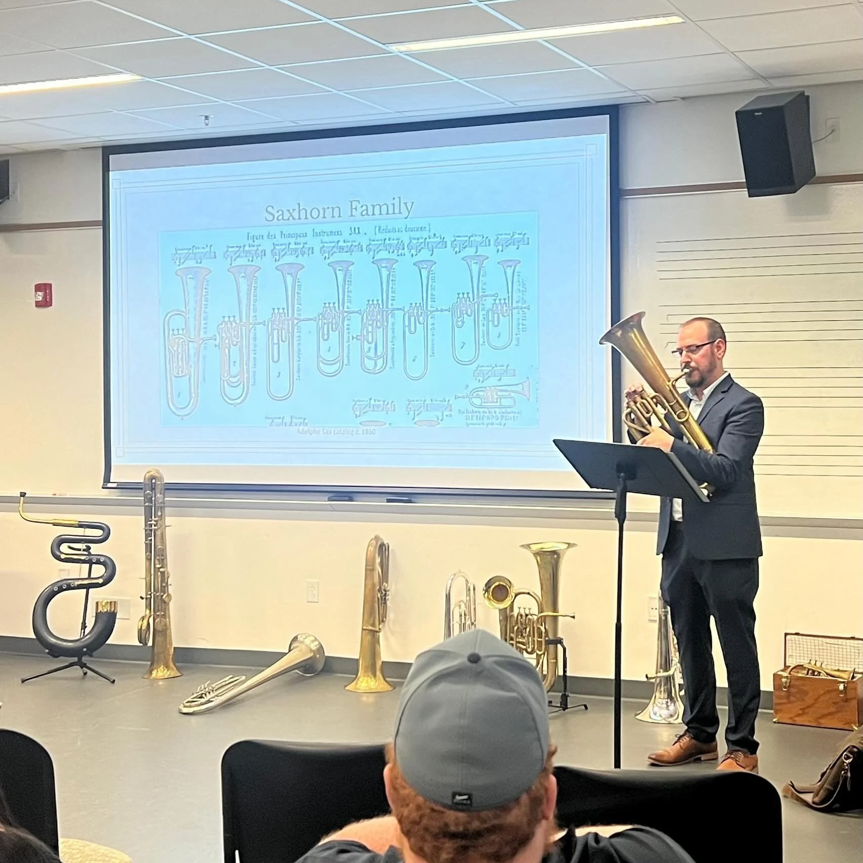 A man in a suit playing a tuba on a stage in front of an audience, with several other brass instruments displayed on the floor and a presentation slide titled 'Saxhorn Family' projected behind him.