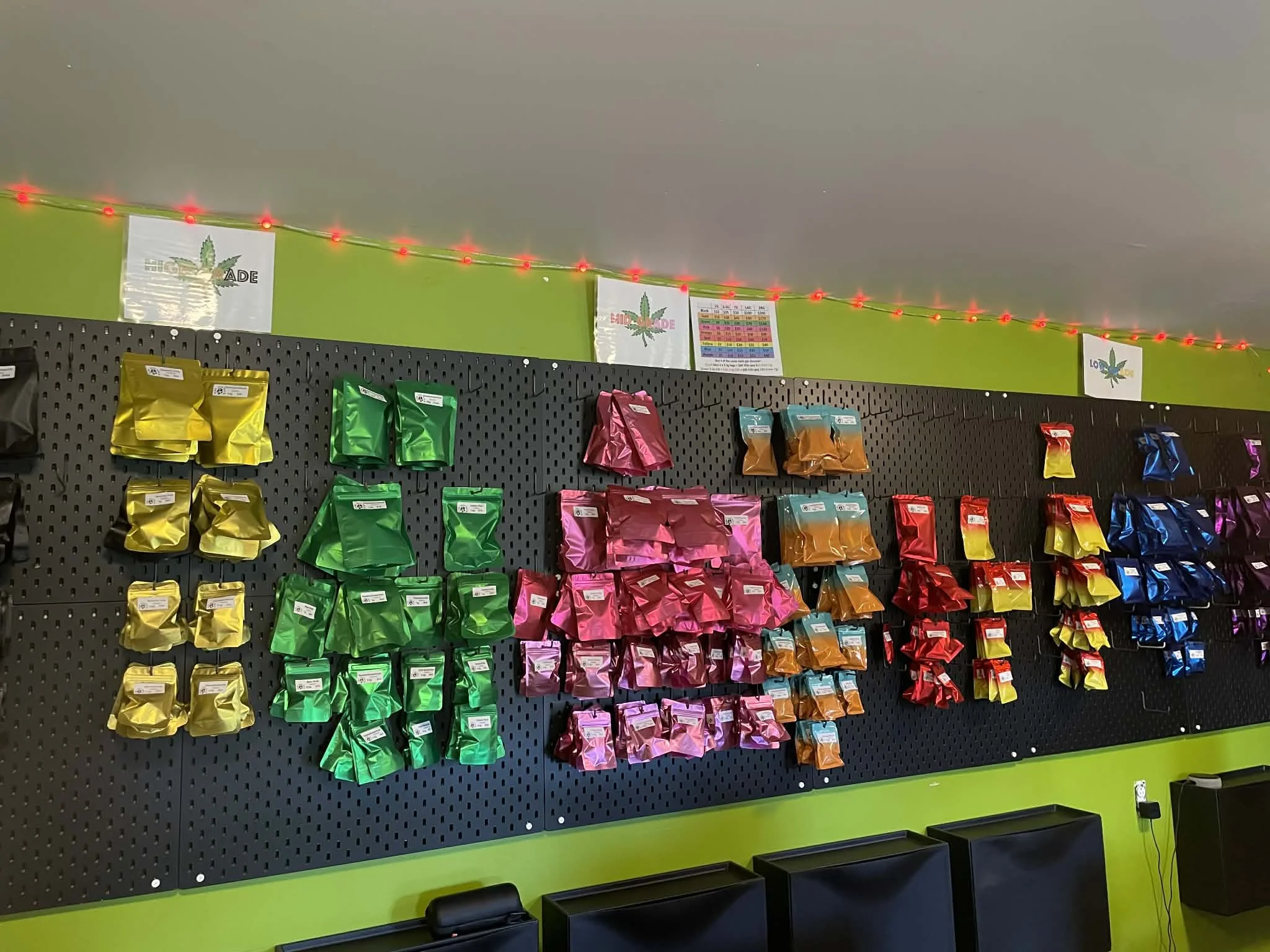 Colorful display of packaged products hanging on a black pegboard in a store, with signs and red string lights above, with a green wall background.