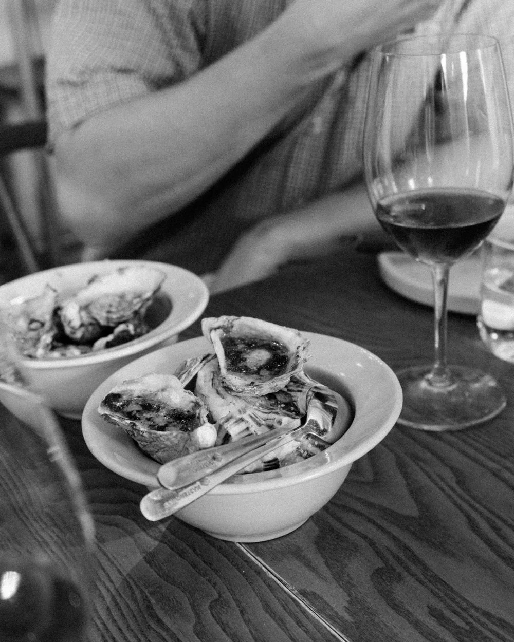 A hand reaching across a table for two bowls of oysters, a glass of wine beside them