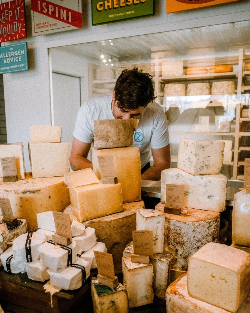 A cheesemonger leaning over a counter stacked with wheels and boxes of cheese