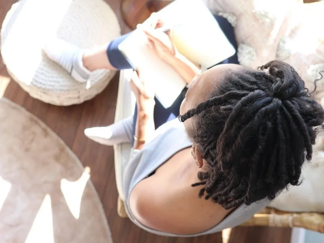 An overhead view of a woman with dreadlocks reading a book, sitting in a cozy setting with a child sitting in front of her, partly visible.