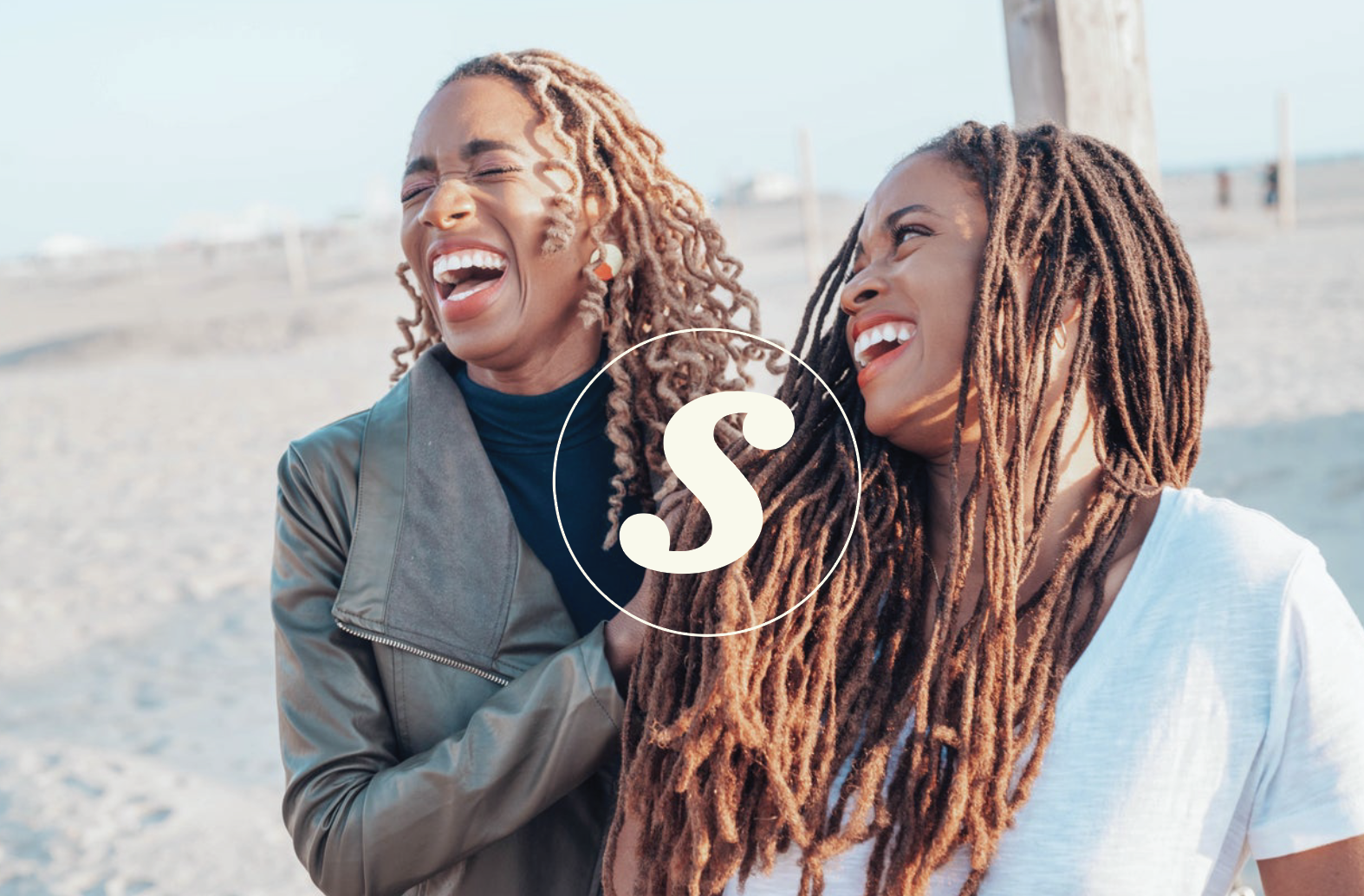 Two women with dreadlocks smiling and laughing on the beach, with a wooden post behind them and a blurred background of sand and sky.