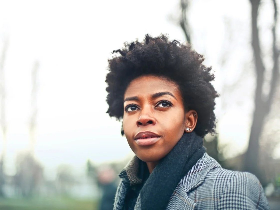 A young woman with natural curly hair looking off into the distance outdoors, wearing a checkered jacket and a dark scarf, with a blurred background of trees.