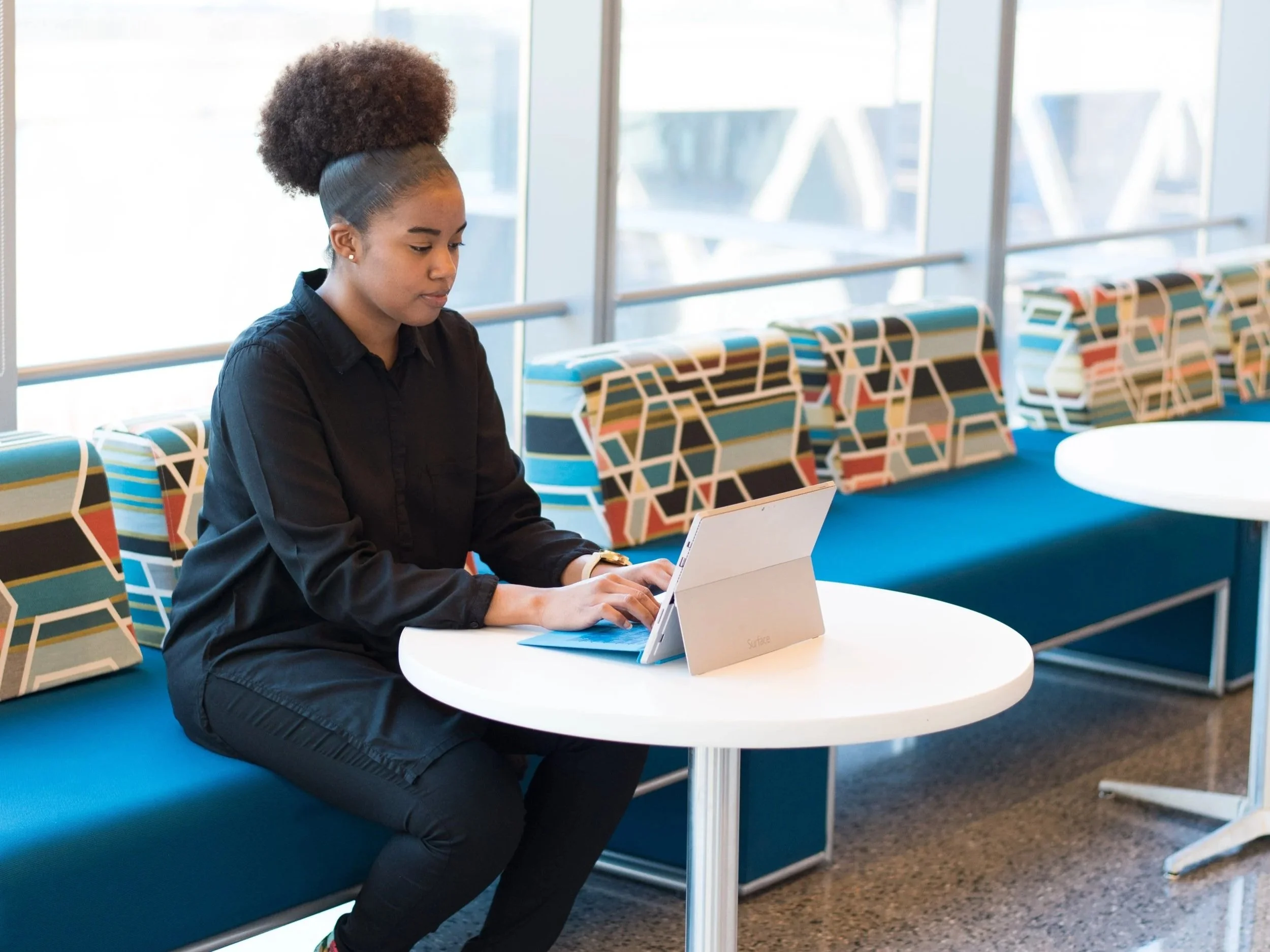 A young woman with curly hair styled in a high puff, wearing a black shirt, sitting at a round white table with a white tablet and a gold watch, in a modern, colorful office or lounge area with patterned chairs and large windows.