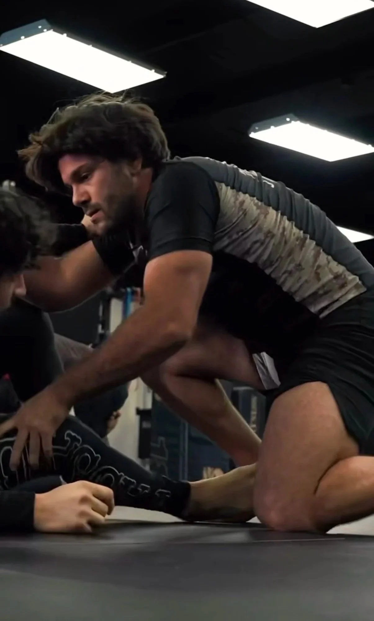 Two people engaging in Brazilian Jiu-Jitsu training on a black mat indoors, with one person in a dominant position on top of the other.