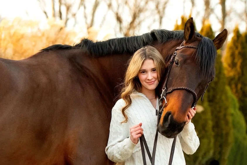Young woman with long blonde hair smiling, standing next to a brown horse outdoors during sunset.