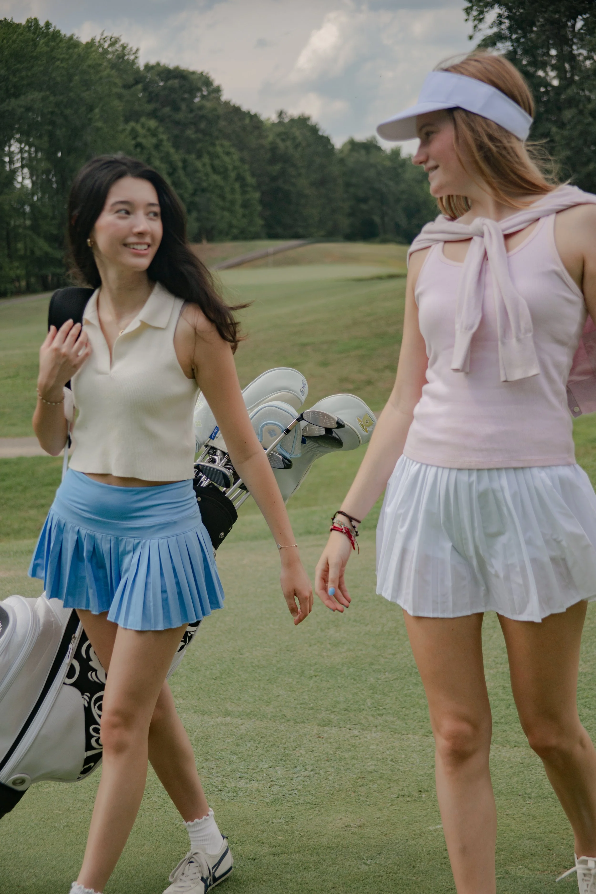 Two young women walk on a golf course. One has dark hair, a sleeveless beige top, and a blue pleated skirt, carrying a golf bag. The other has light hair, a pink sleeveless top with a tied collar, and a white pleated skirt, wearing a visor. They are smiling and looking at each other, with trees and a cloudy sky in the background.