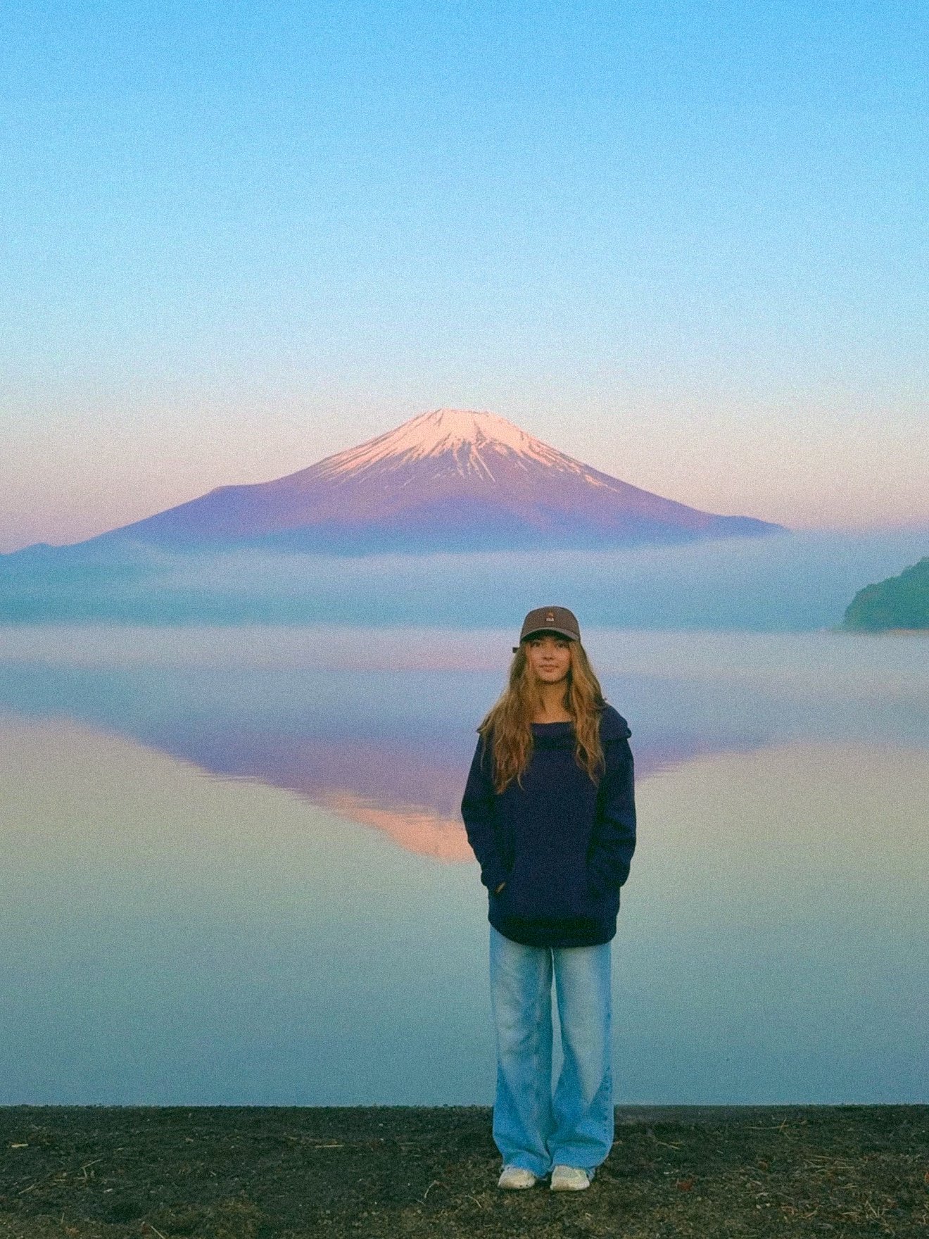 A woman standing in front of a calm lake with Mount Fuji in the background, snow-capped and partly obscured by mist in soft pastel colors during dawn or dusk.