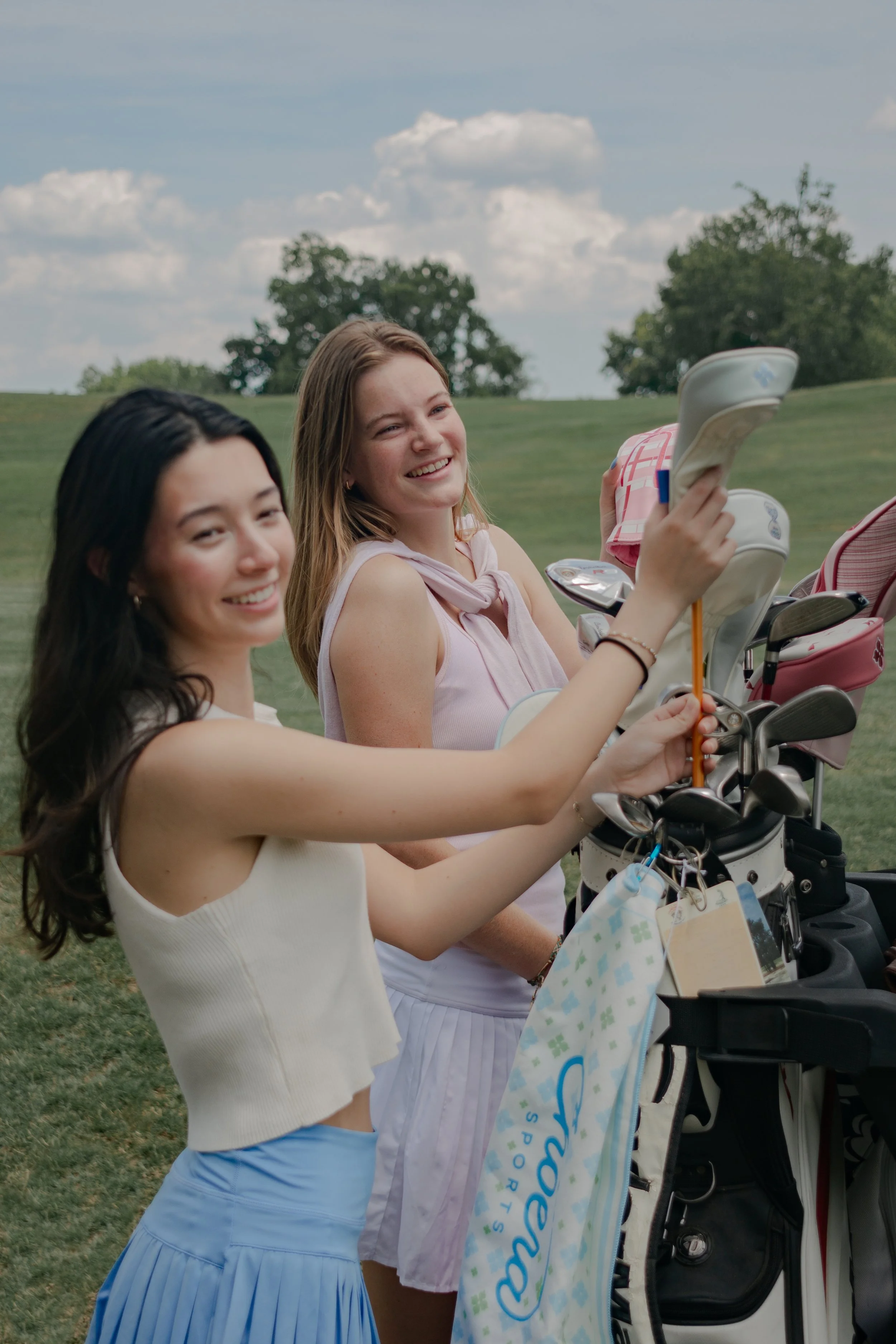 Two young women on a golf course near a golf cart. One woman is adjusting a golf club in the golf bag, and the other is holding a golf tee. They are smiling and enjoying a sunny day.