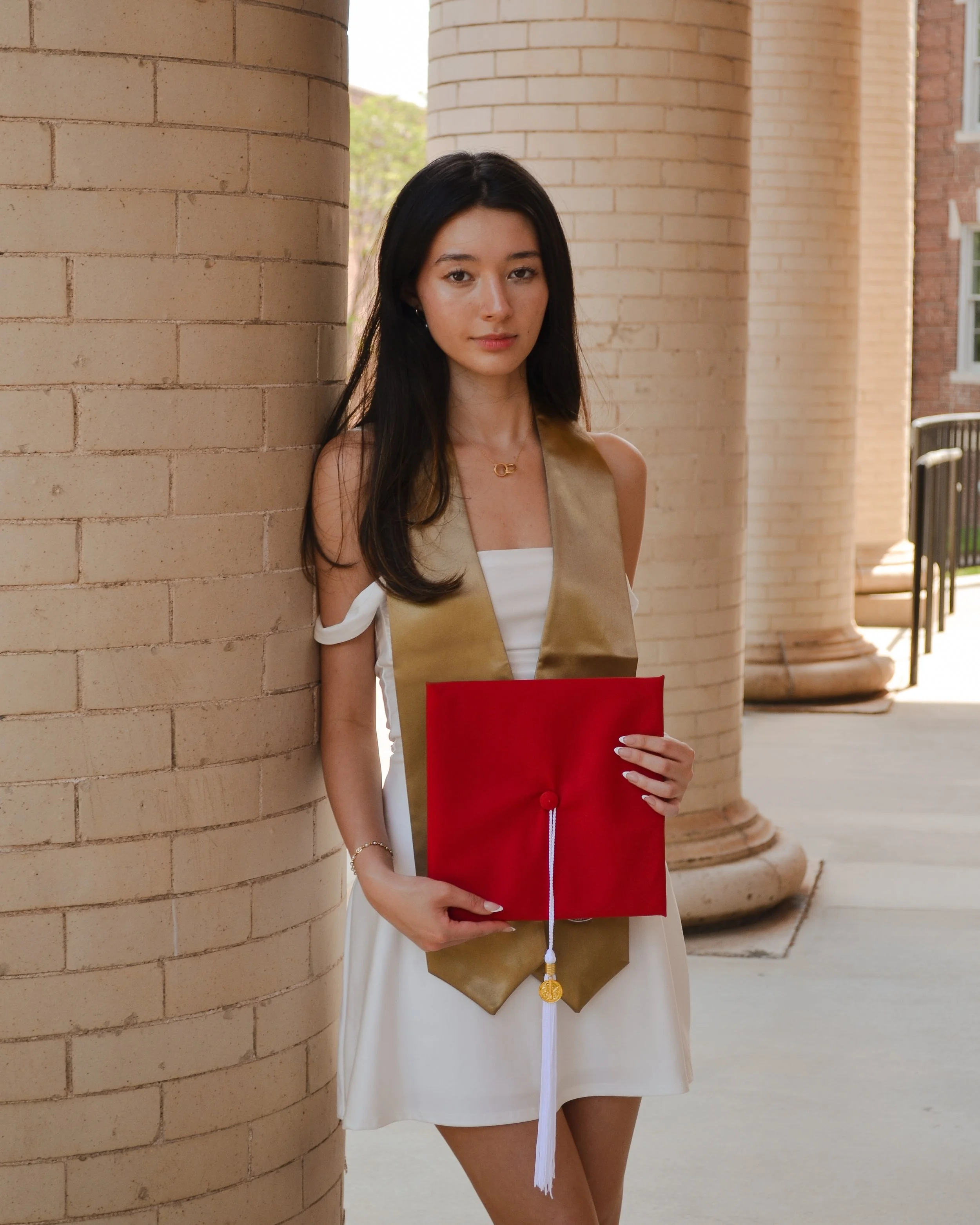 A young woman with long dark hair, wearing a white dress and a gold necklace, stands outdoors near brick columns, holding a red graduation cap.