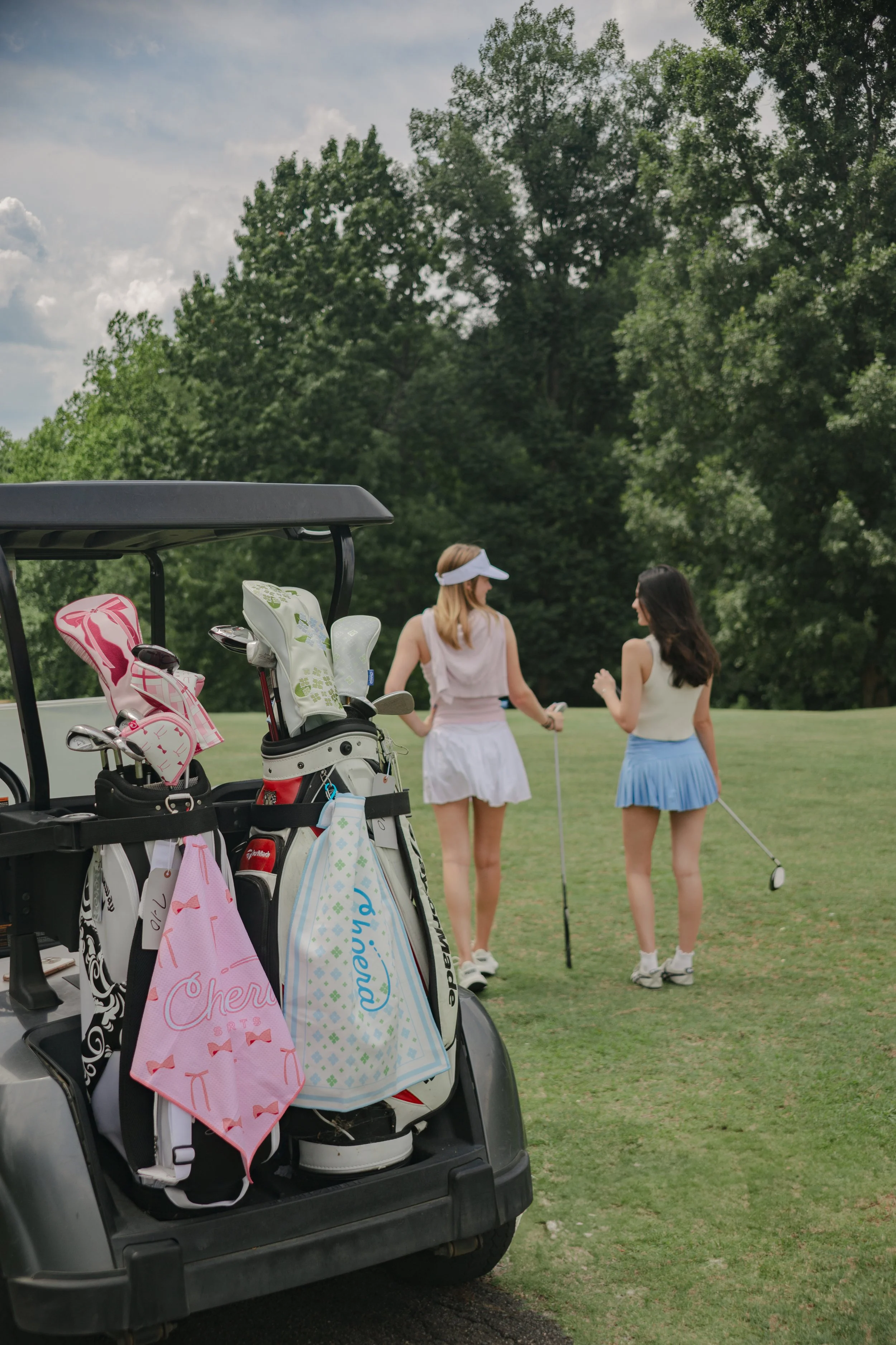 Golf cart with golf bags and towels on the back, parked on a golf course, with two women playing golf in the background under a partly cloudy sky.
