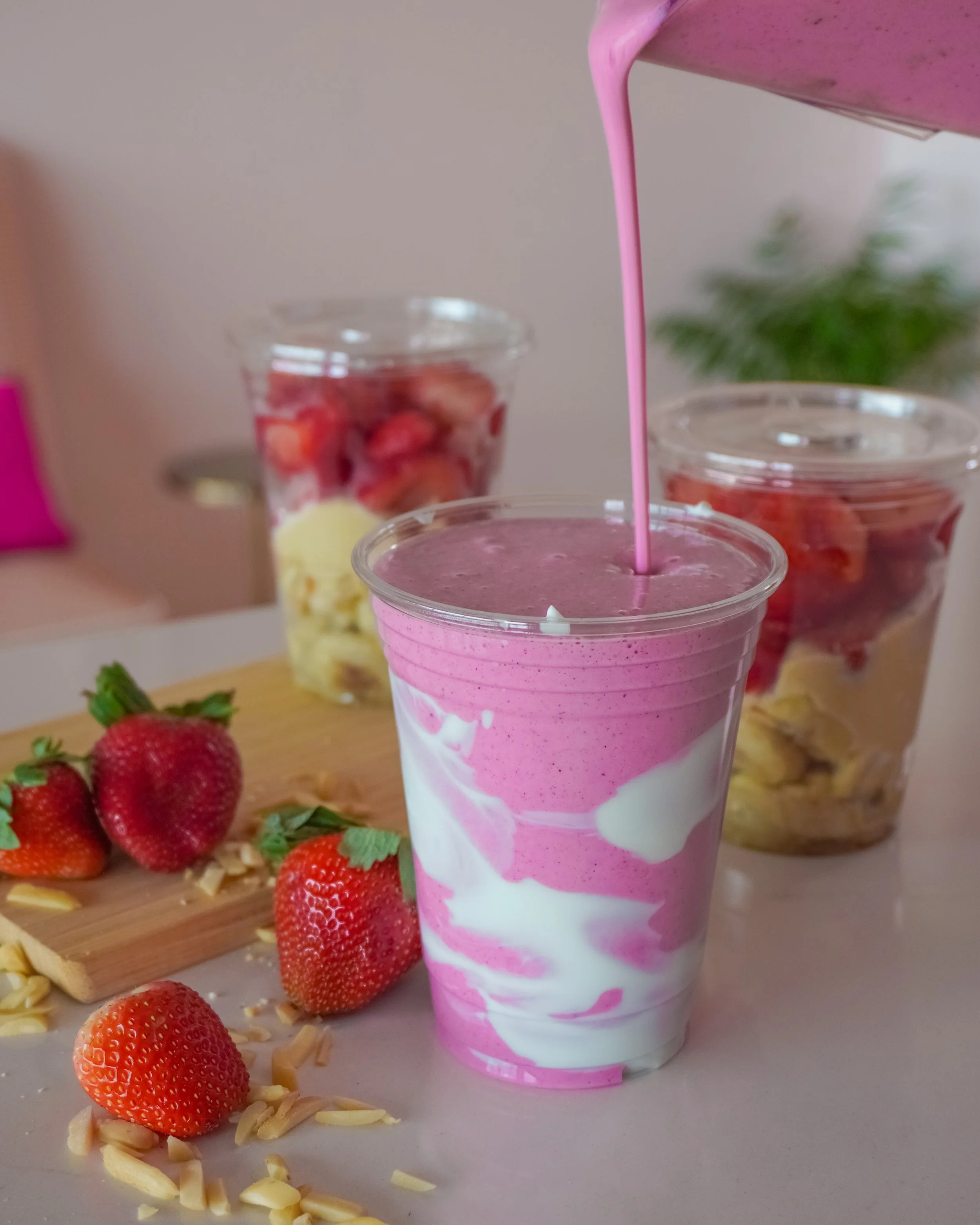 A strawberry smoothie in a paper cup with pink and white swirl pattern, surrounded by fresh strawberries, chopped almonds, and dessert cups with strawberries and cream in the background.