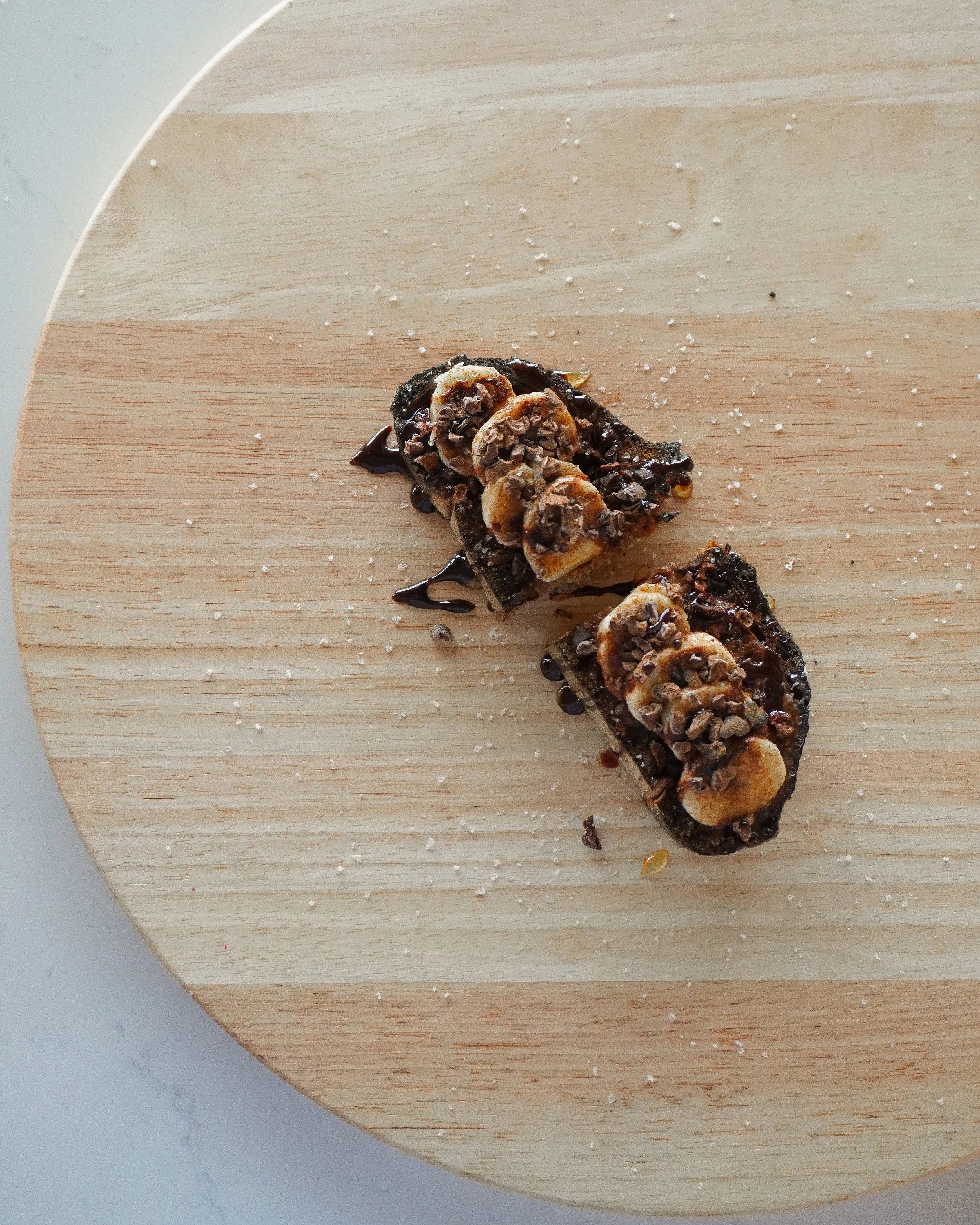 Close-up of a banana and chocolate dessert topped with chopped nuts and drizzled with chocolate syrup on a round wooden surface.