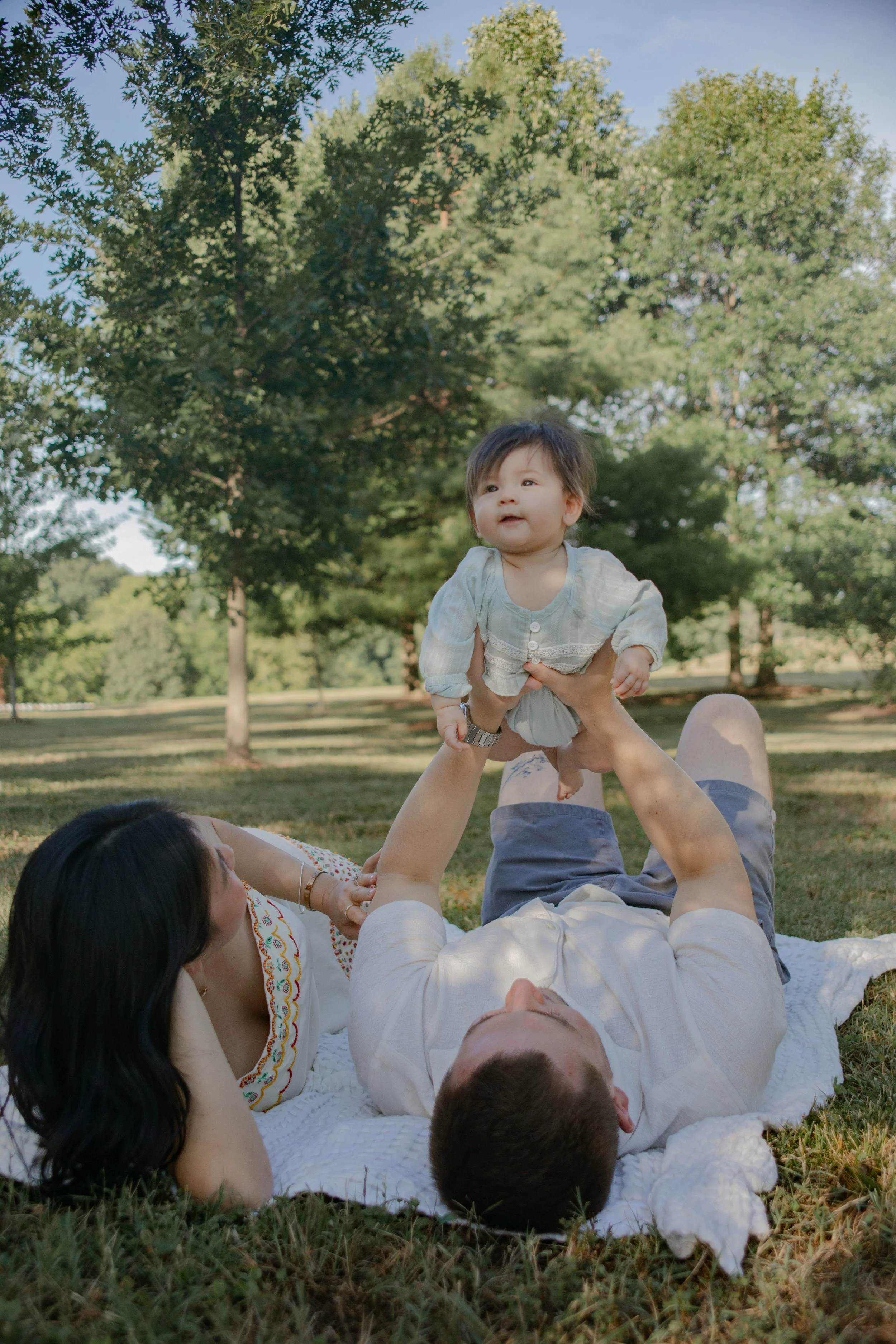 A family creating a playful moment outdoors with a woman lying on the grass, a man lying down, and a child in a light-colored dress being lifted in the air by the man, all surrounded by trees and green grass.