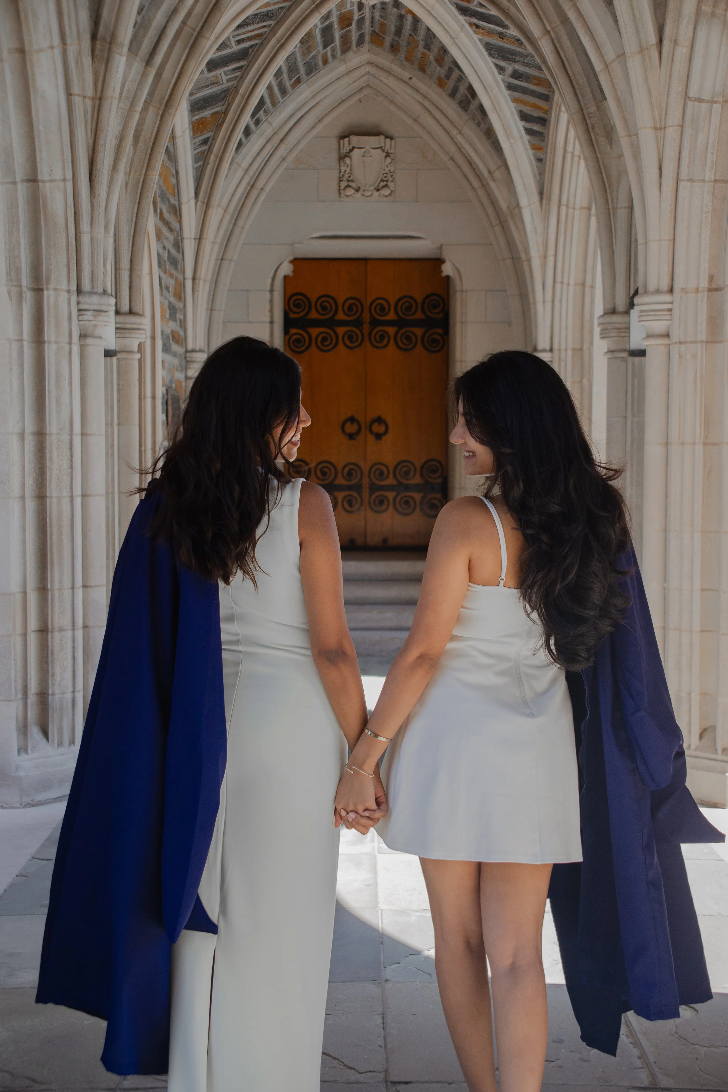 Two women in white dresses holding hands, standing in an arched stone doorway with a wooden door behind them, smiling at each other.