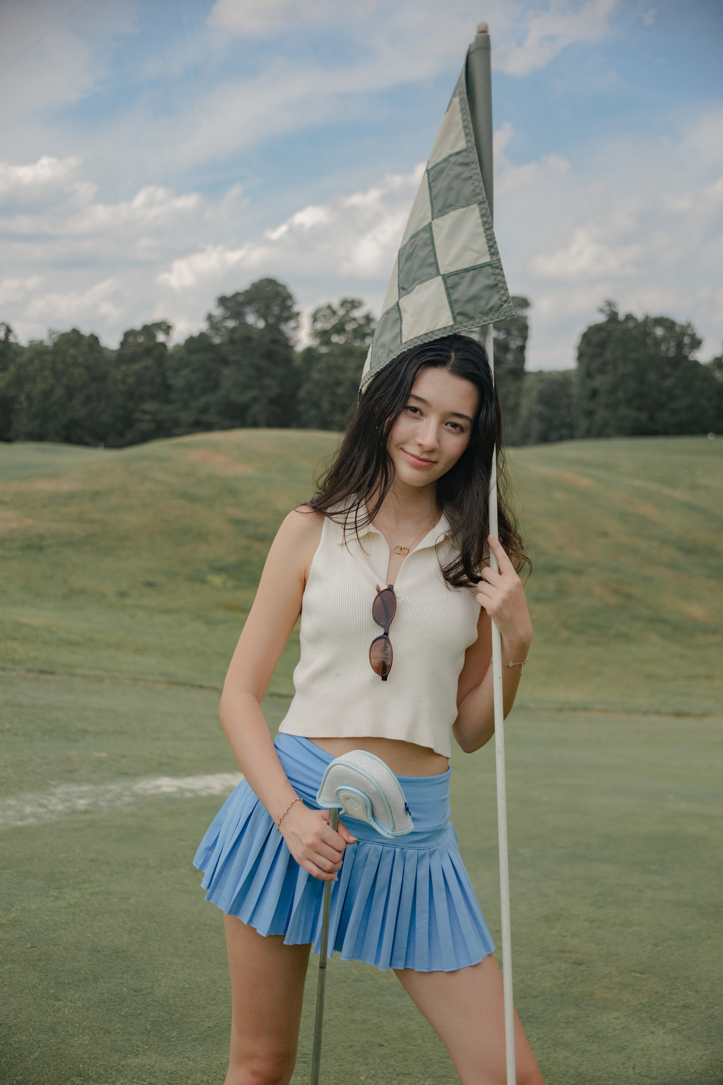 Young woman on a golf course holding a golf club and a golf flag, wearing a white sleeveless top and a blue pleated skirt, with sunglasses hanging from her top and a bracelet, with trees and cloudy sky in the background.
