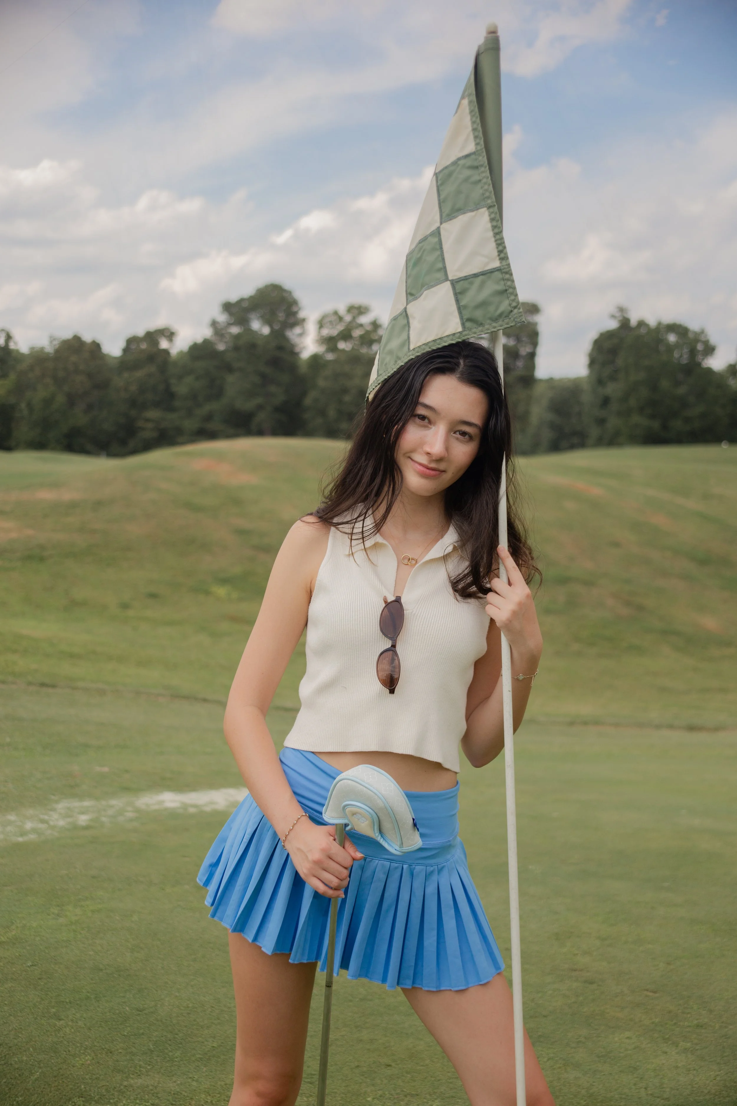 A young woman standing on a golf course holding a golf club and a golf flag; she is wearing a sleeveless top and a pleated blue skirt, with sunglasses hanging from her top.