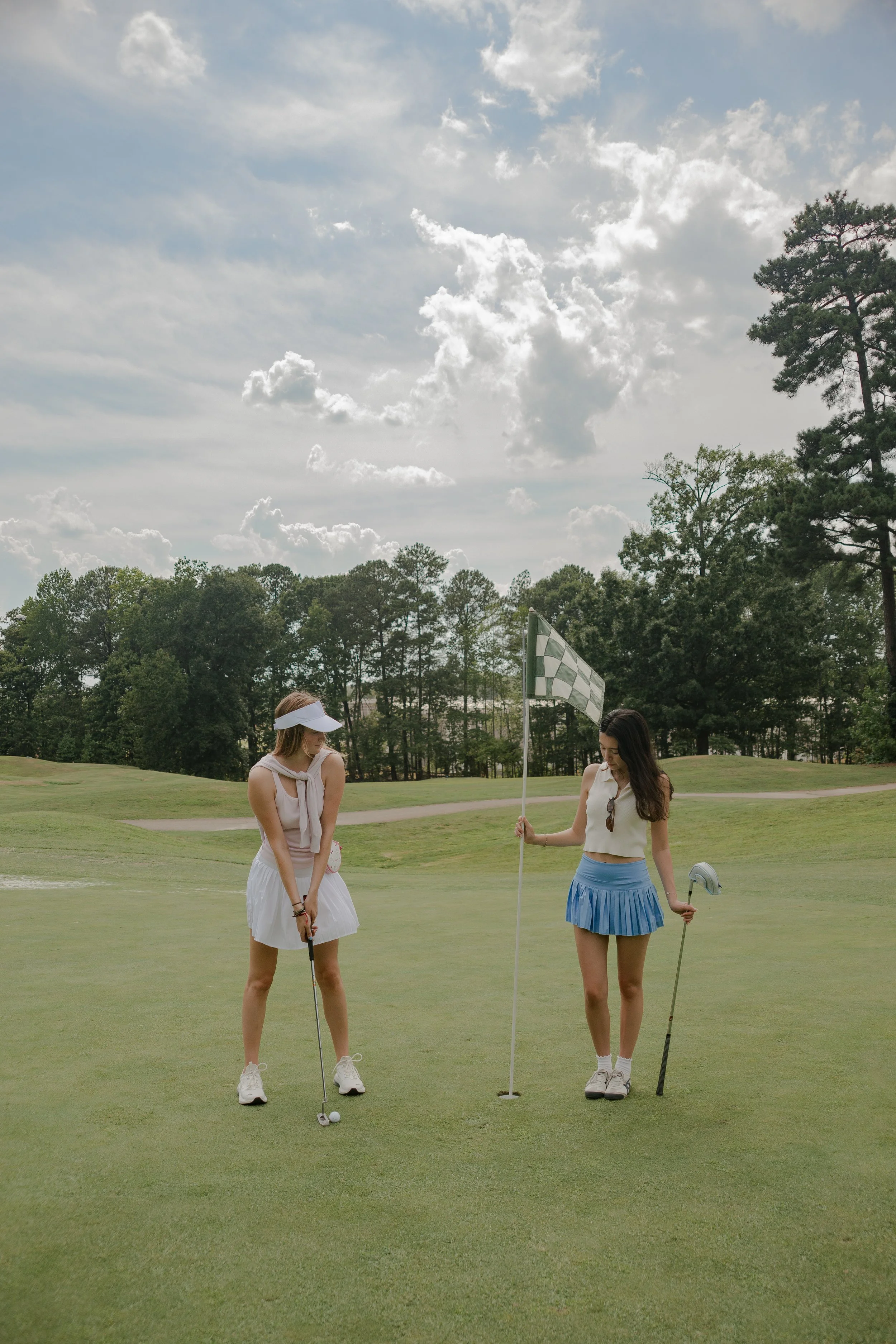 Two young women standing on a golf course near a hole, one with a golf club and the other with a golf flag, under a partly cloudy sky, with trees in the background.