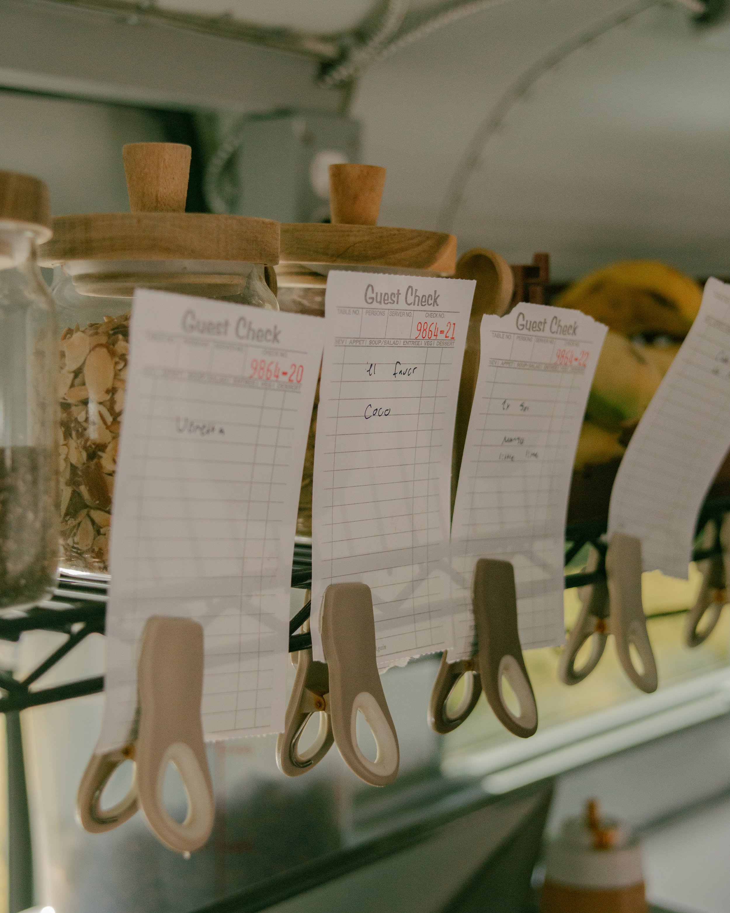 Close-up of small wooden clothespins hanging from a white string with guest check notes attached to each pin, in a kitchen or dining setting.