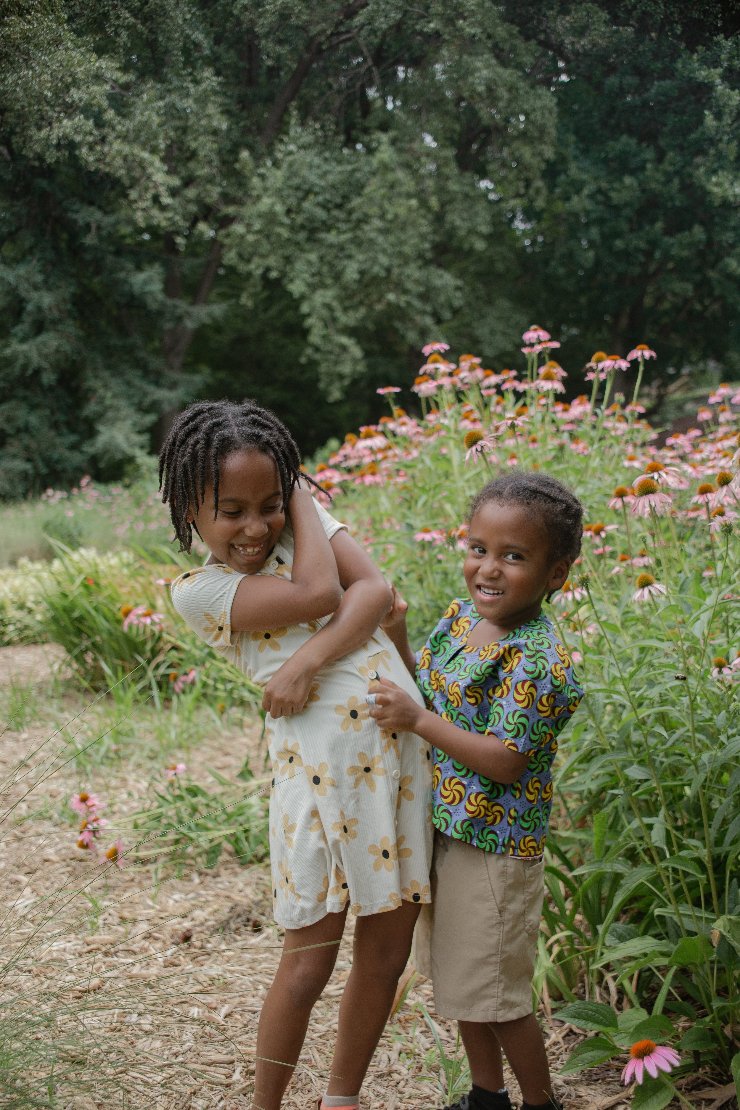 Two children, a girl and a boy, playing in a garden with pink flowers and green foliage.