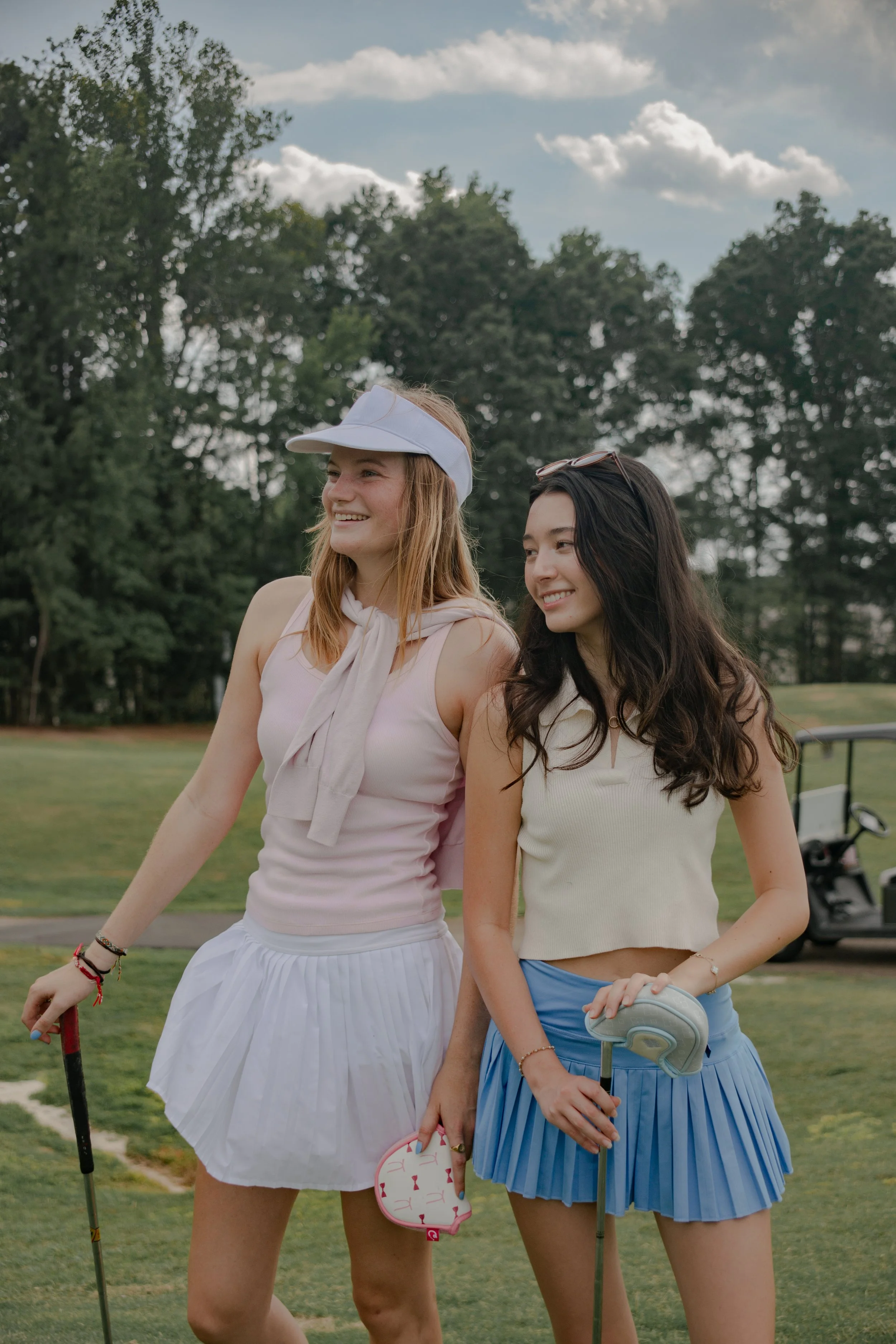 Two young women standing on a golf course, smiling and holding golf clubs, with trees and a partly cloudy sky in the background.