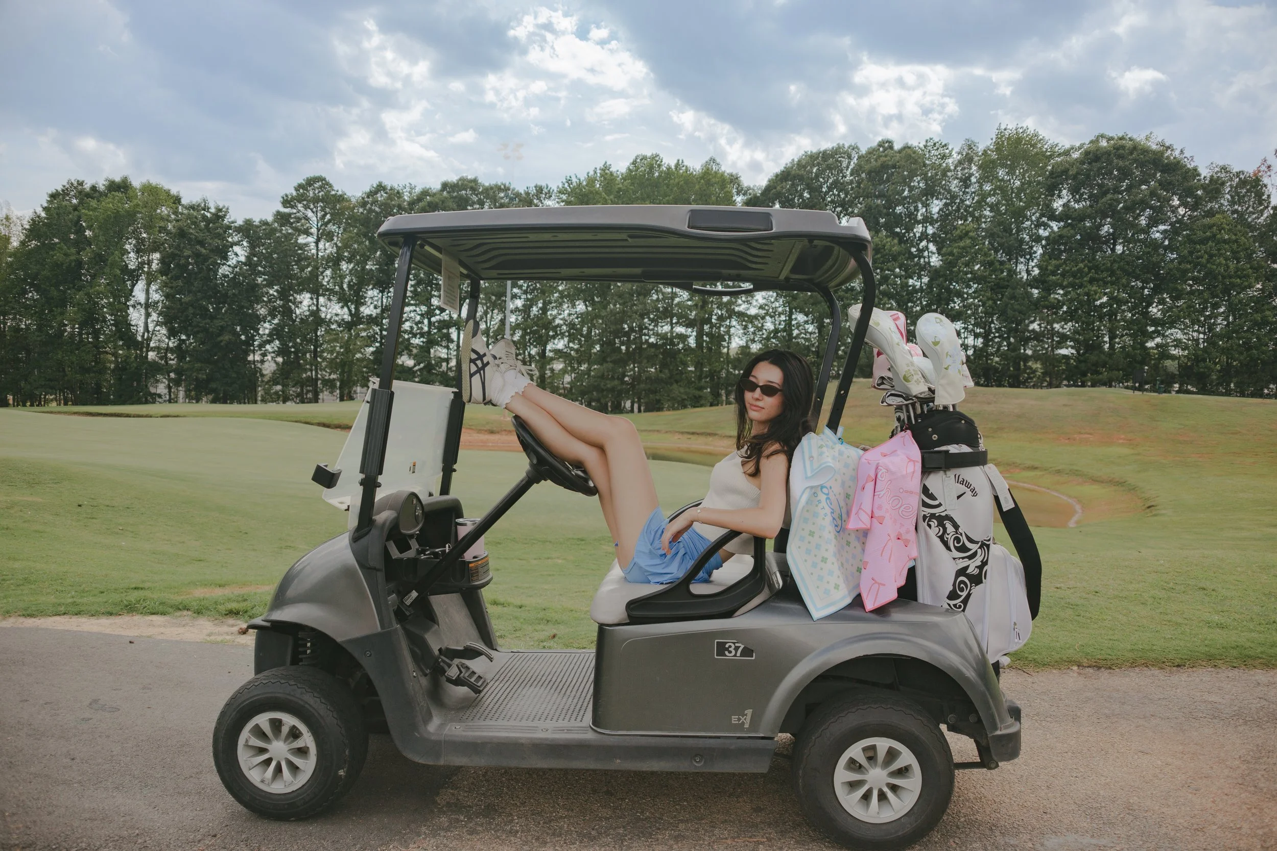 A woman sitting with her legs stretched out in a golf cart on a golf course, with golf bags and clubs on the back. She is wearing sunglasses, a white top, and blue shorts, and has dark hair. The sky is partly cloudy, and there are trees in the background.