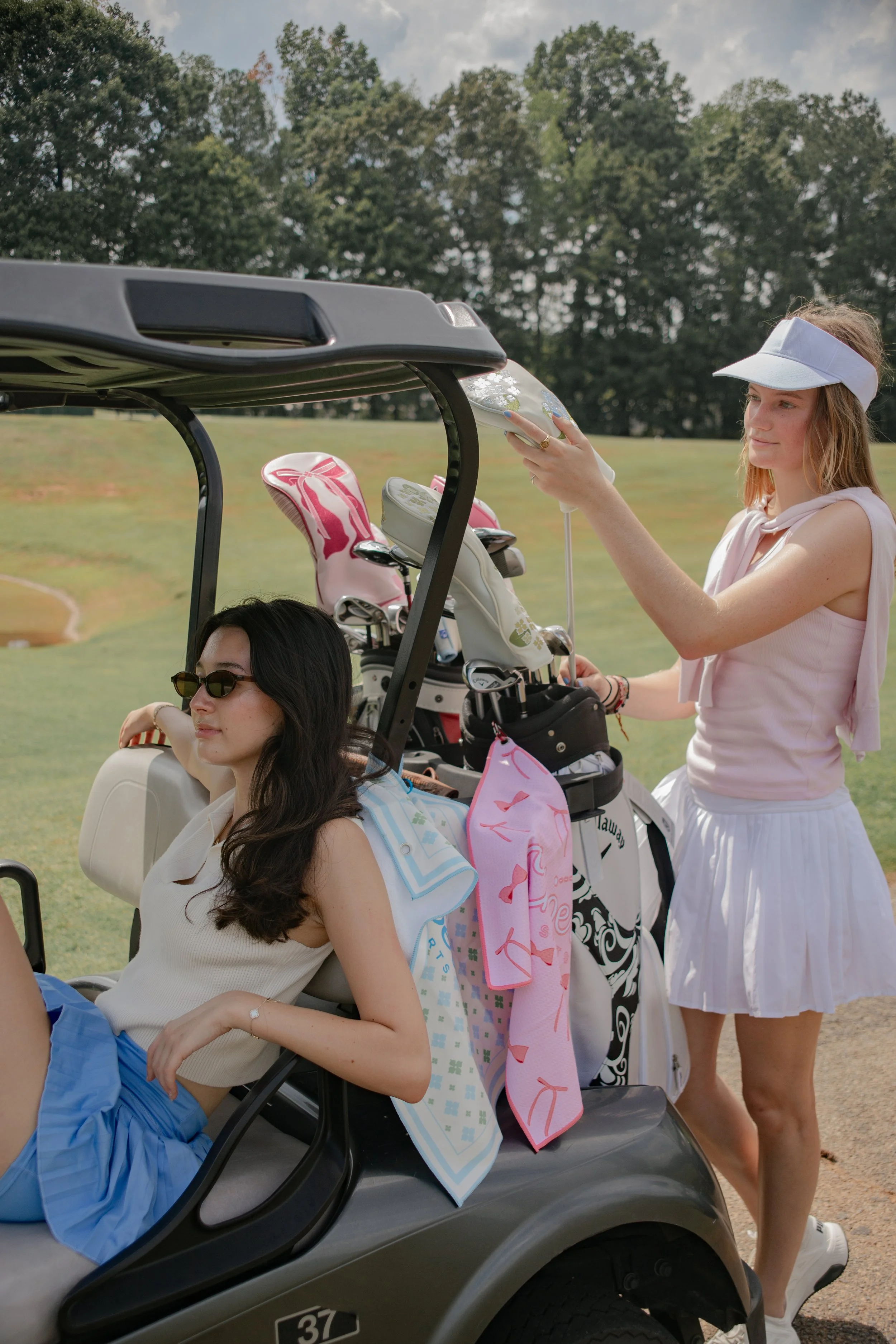 Two women are on a golf course beside a golf cart. One woman with dark hair and sunglasses is seated in the golf cart, while the other woman with light hair, wearing a white visor, white sleeveless top, and white skirt, is standing outside the golf cart, holding a shiny silver object, possibly a golf ball dispenser. Golf clubs are visible in the cart, and a pink towel and other items are hanging from the cart. The background shows a grassy field and trees.