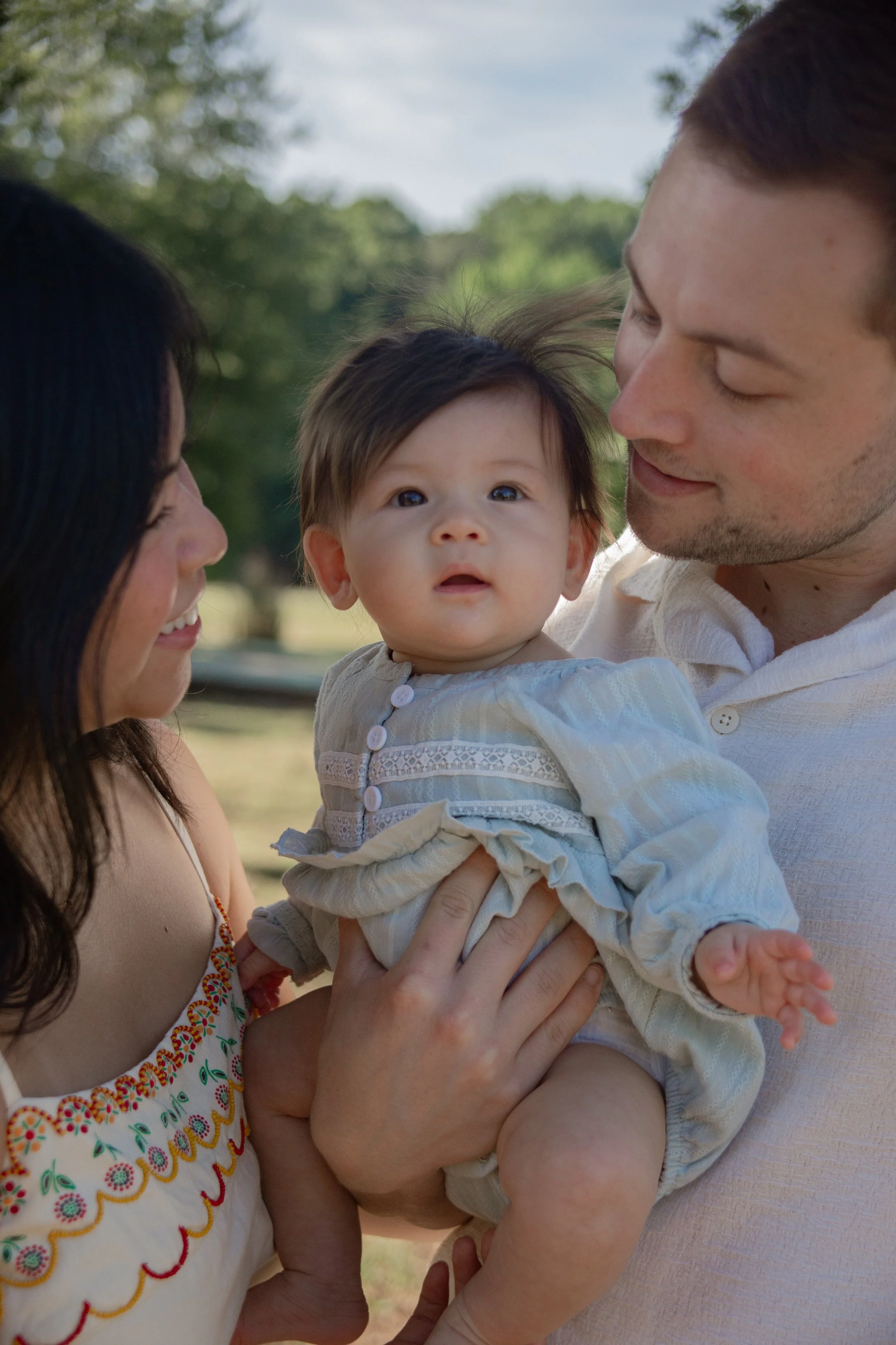A family outdoors, with a woman on the left smiling, a man on the right holding a baby, and a background of green trees and a cloudy sky.